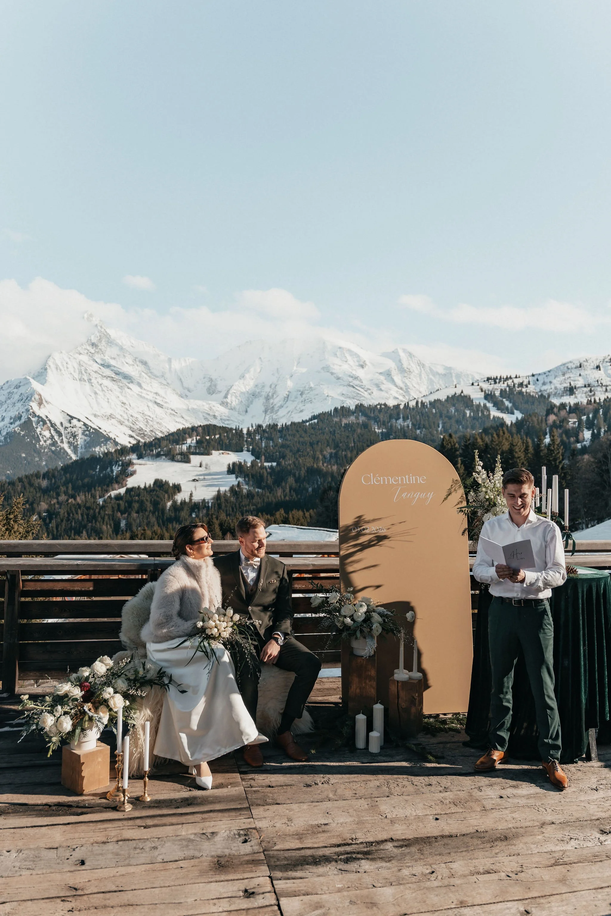 Un couple de mariés assis lors d'une cérémonie en plein air avec une montagne enneigée en arrière-plan. La mariée porte une robe blanche et un manteau en fourrure, et le marié porte un costume gris. Un jeune homme debout à droite, probablement le cél