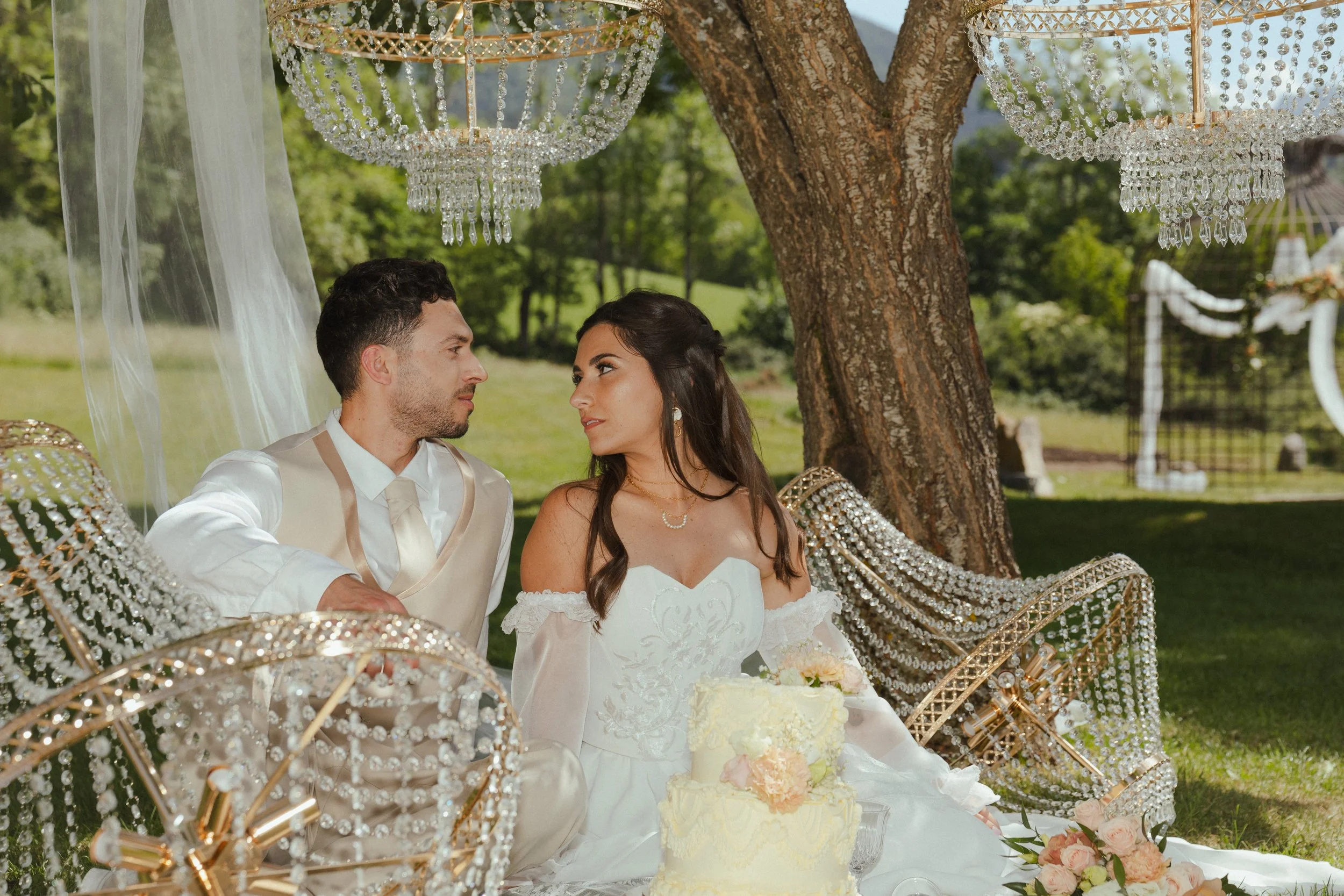 Un couple de jeunes mariés assis sous un arbre lors d'un mariage en extérieur, entouré de décorations en cristal et de lampions décoratifs.