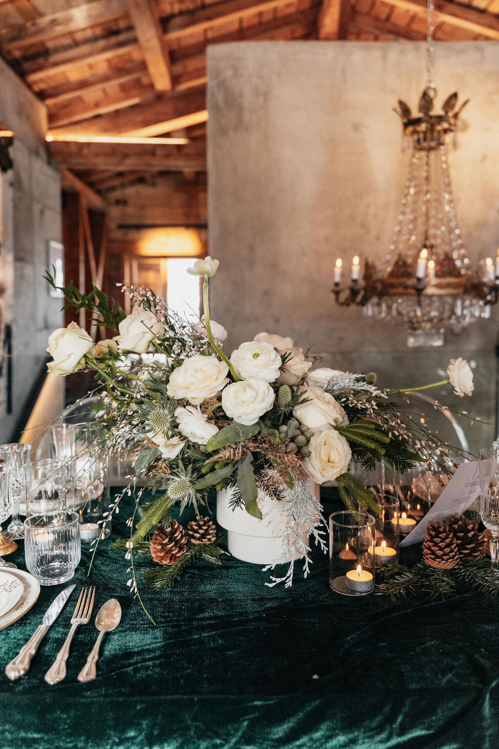 Une décoration de table de mariage avec un grand bouquet de fleurs blanches, des bougies dans des petits récipients en verre, et des pommes de pin, sur une nappe en velours vert foncé, dans une salle avec un plafond en bois et un chandelier en crista