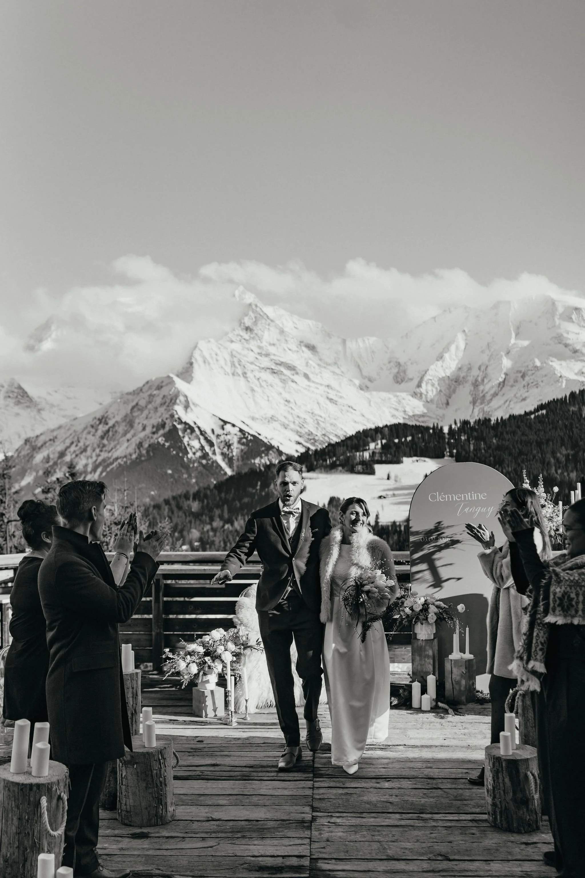 Mariage en plein air avec montagnes enneigées en arrière-plan, couple de mariés avec invités applaudissant, décor floral et bougies, panneau avec le nom Clémentine Tanguy.