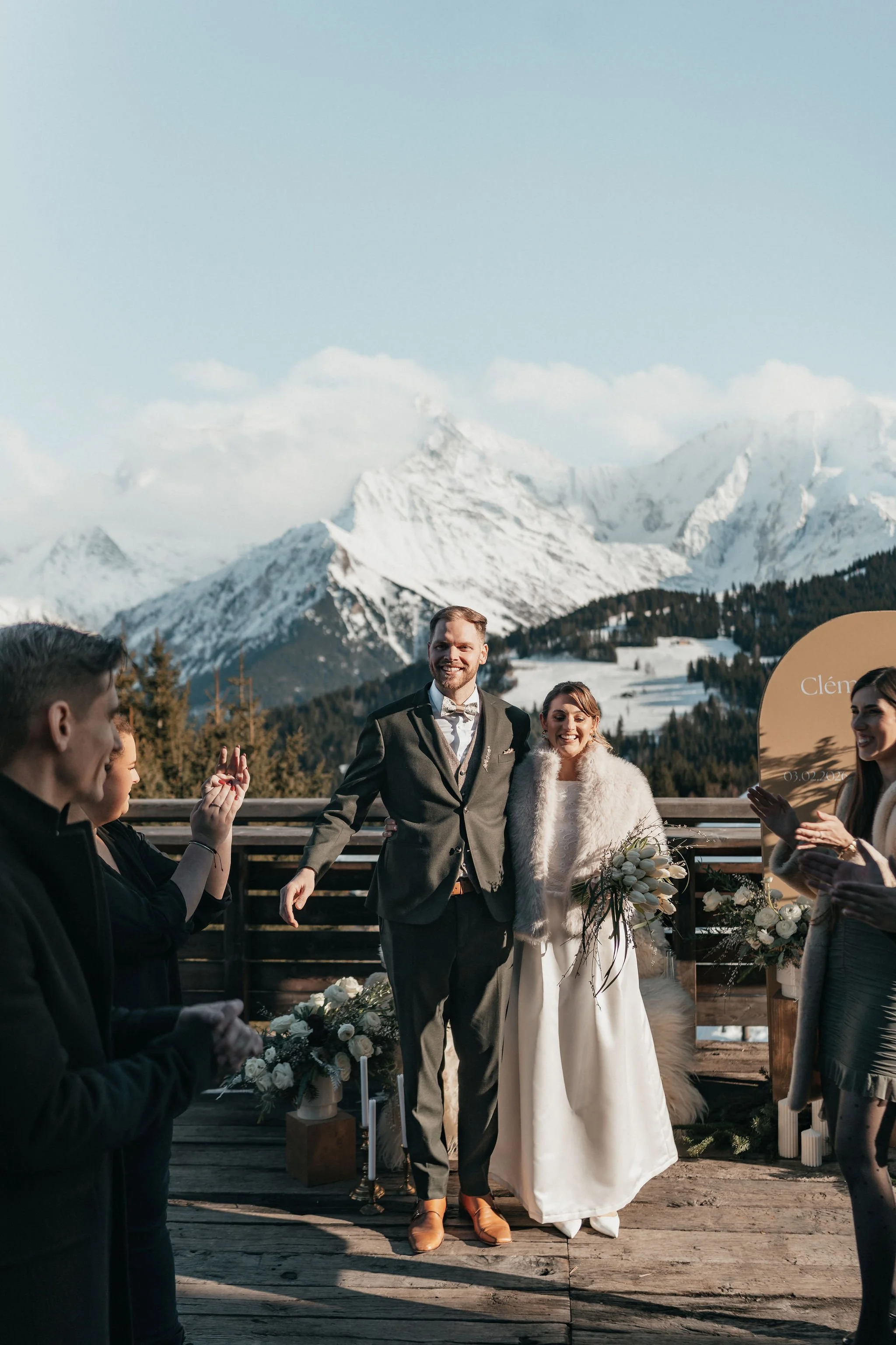 Un couple de mariés heureux lors de leur mariage en montagne, entouré de leurs amis, avec un décor de montagnes enneigées en arrière-plan.