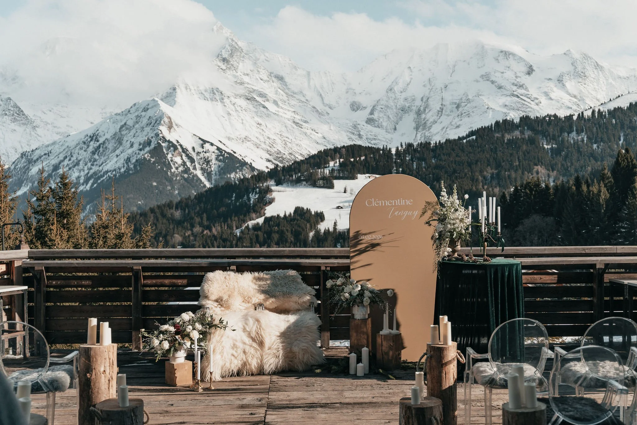 Décoration d'un lieu en plein air avec une vue sur des montagnes enneigées. Il y a un fauteuil avec une couverture pelucheuse, des fleurs, des bougies, et une grande carte avec le nom 'Clémentine Tanguy' et la date 15.02.2024.