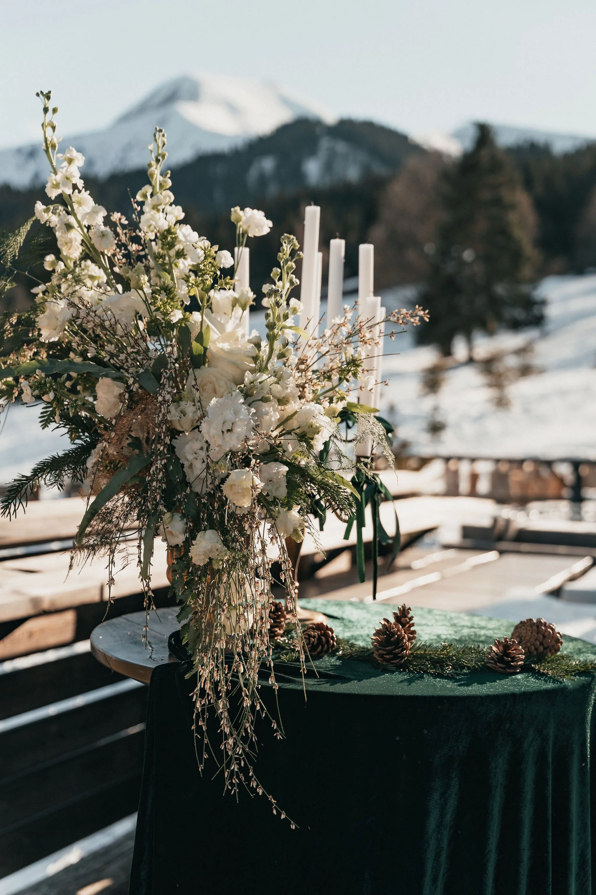 Décoration florale blanche avec des bougies sur une table couverte d'un linge vert, dans un décor de montagnes enneigées et de forêts.