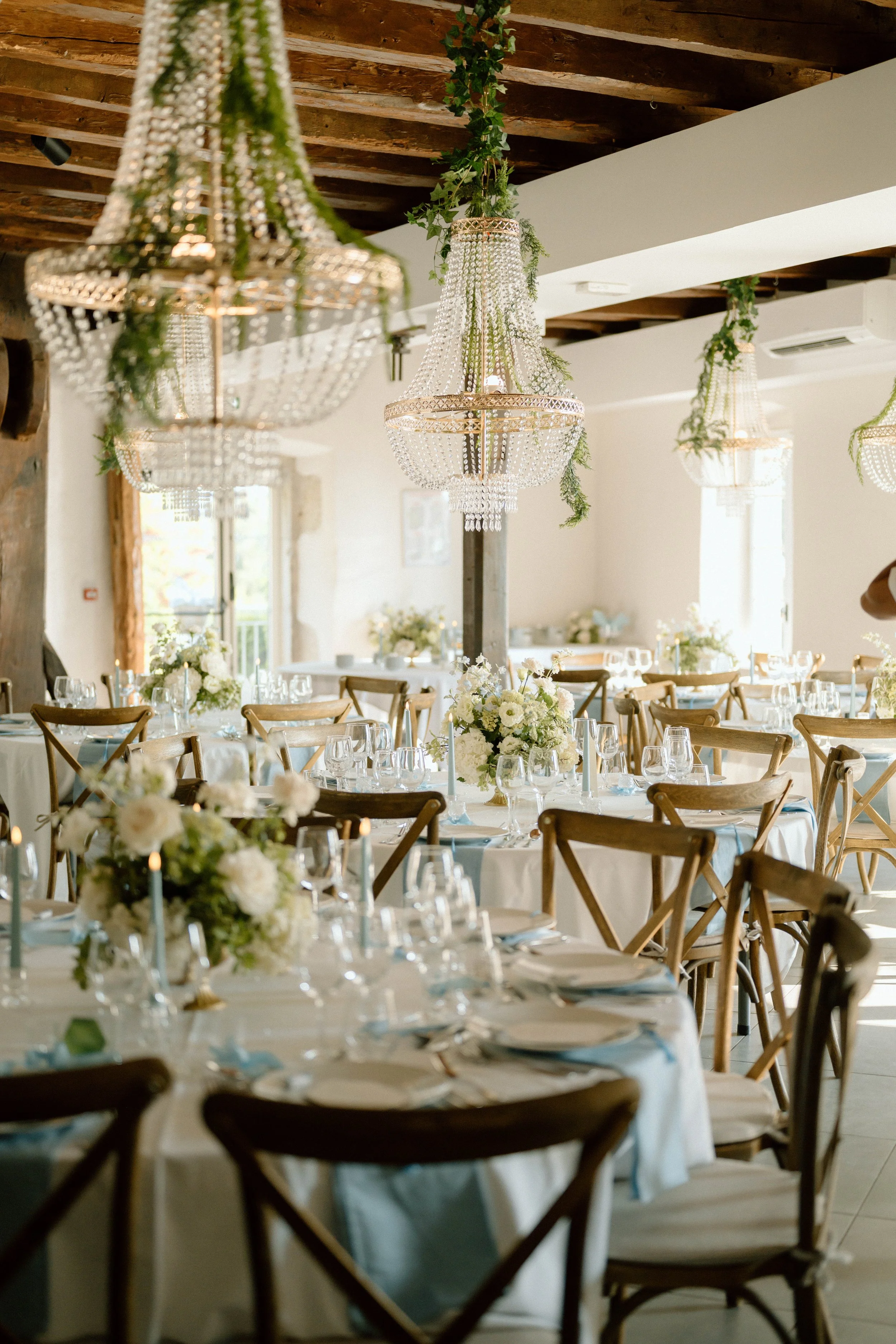 Salle de réception décorée avec des chandeliers en cristal suspendus au plafond, tables rondes avec nappes blanches, assiettes, couverts, verres à vin et arrangements floraux blancs et verts.