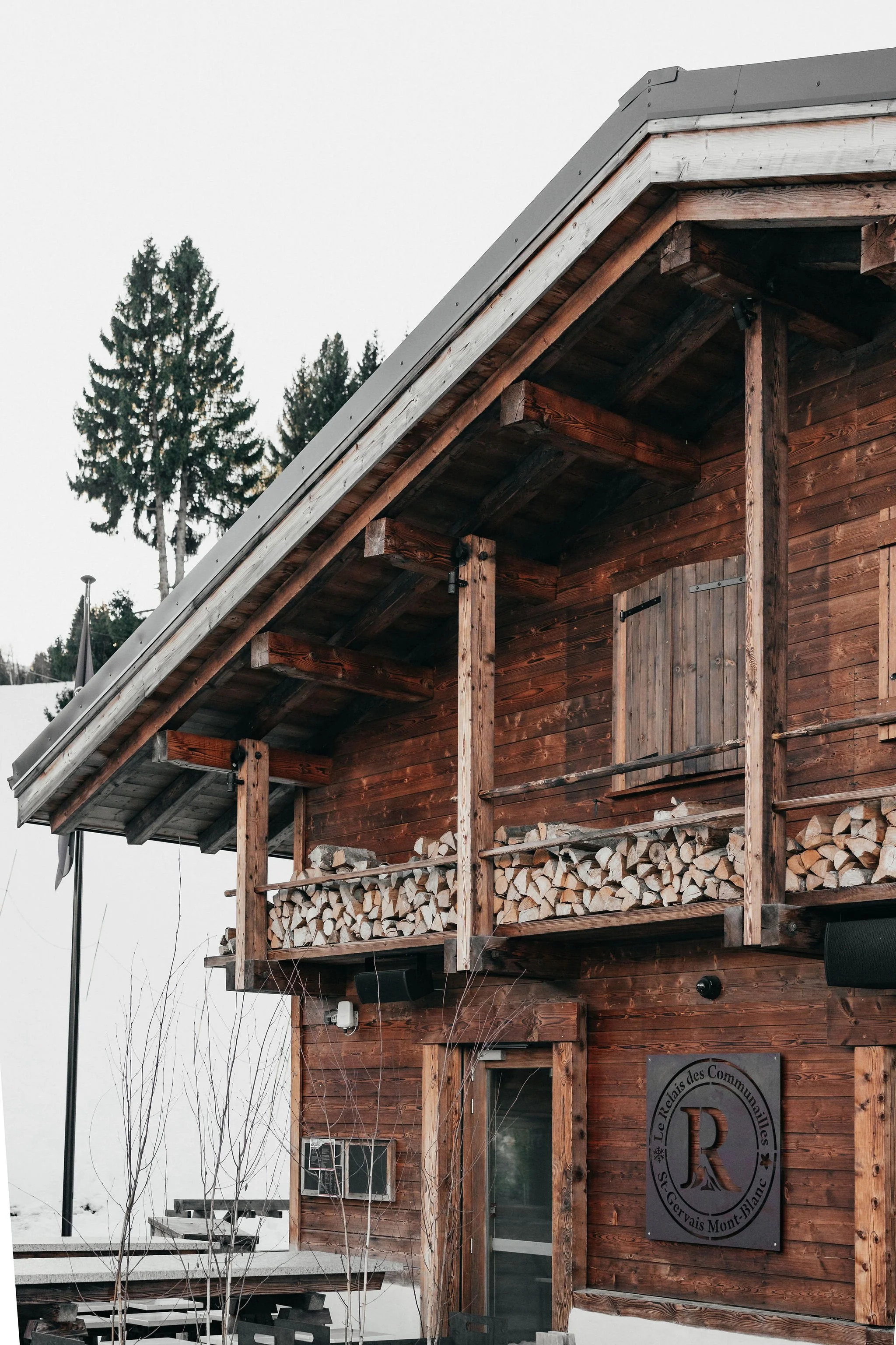 Une cabine en bois avec un balcon rempli de bois de chauffage, entourée de neige, dans un environnement enneigé avec des arbres en arrière-plan.