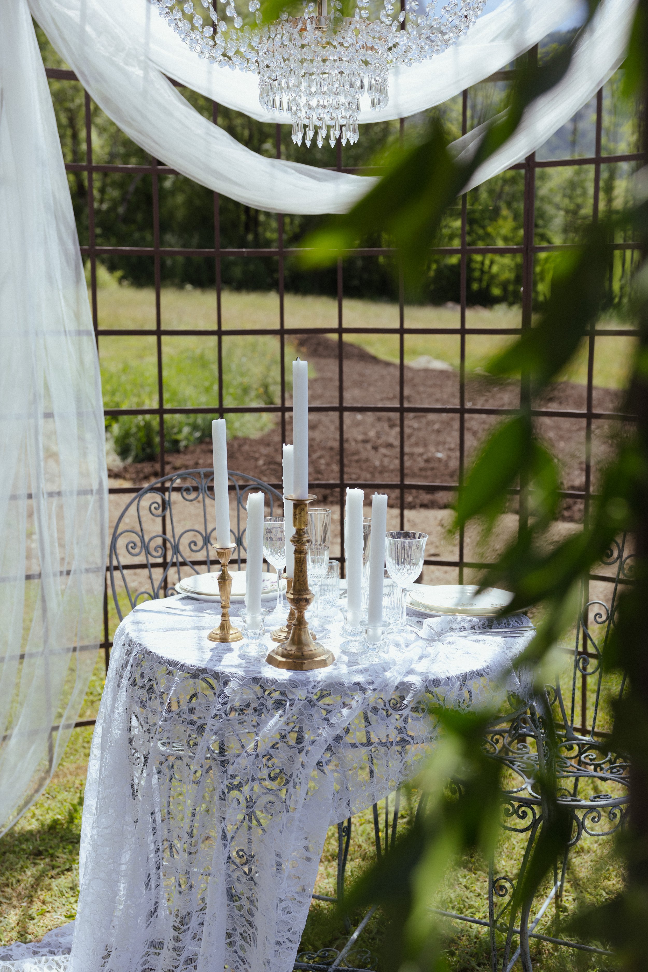 Table élégante décorée avec des chandeliers en or et des bougies blanches, entourée de chaises en fer forgé, dans un cadre extérieur avec une grille en métal et des draps blancs suspendus, entouré de verdure