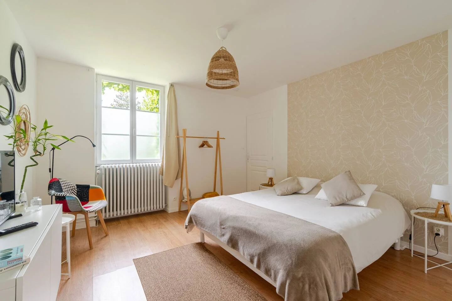 Chambre moderne avec un lit king-size, murs blancs, un mur accent avec un motif floral beige, une lampe suspendue en osier, fenêtres avec rideaux clairs, mobilier en bois clair, un fauteuil coloré, un porte-vêtements en bois, une table de chevet avec lampe, un tapis beige.