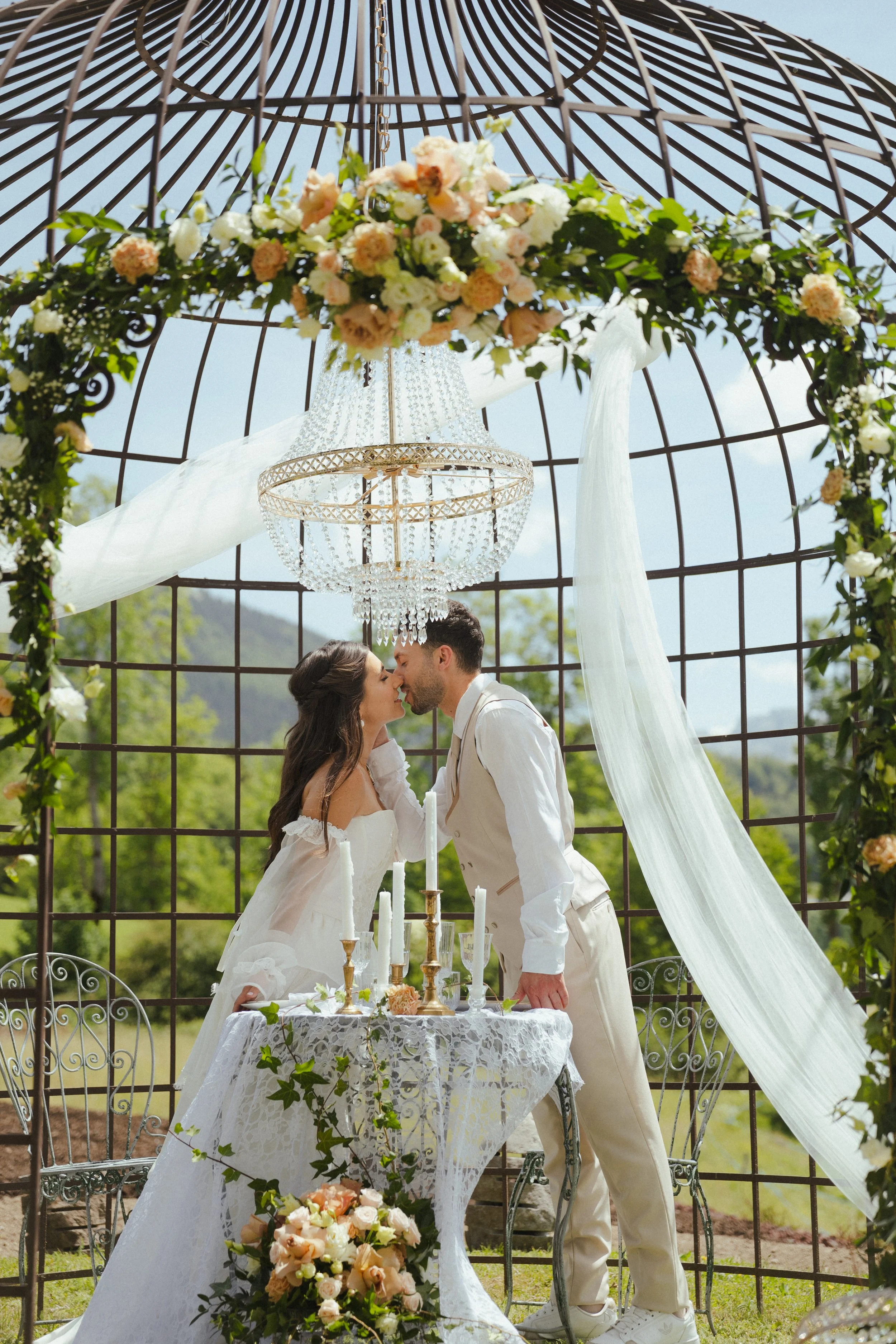 Un couple en tenue de mariage s'embrasse sous une arche décorée de fleurs lors d'une cérémonie en plein air, avec une sculpture en métal en arrière-plan et une table ornée de bougies et de fleurs.
