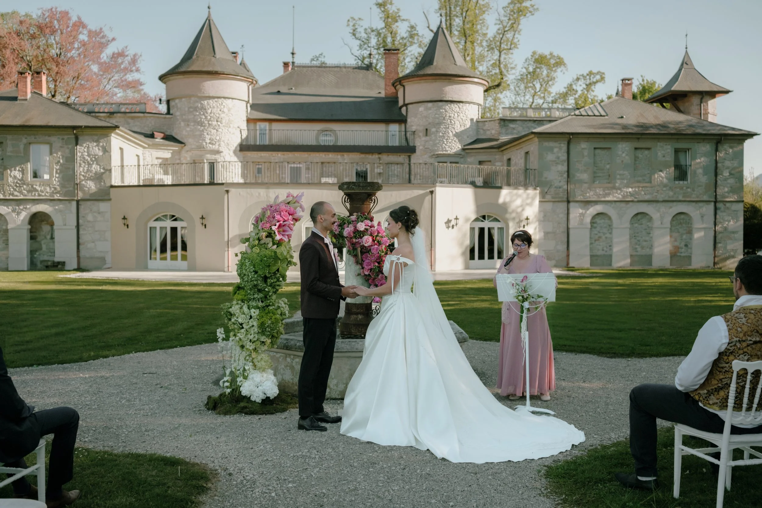 Un couple se tenant la main lors d'une cérémonie de mariage en plein air devant un château en pierre avec des tours. La mariée porte une robe blanche et un voile, le marié un costume sombre. Une femme avec des lunettes et une robe rose pale lit un di