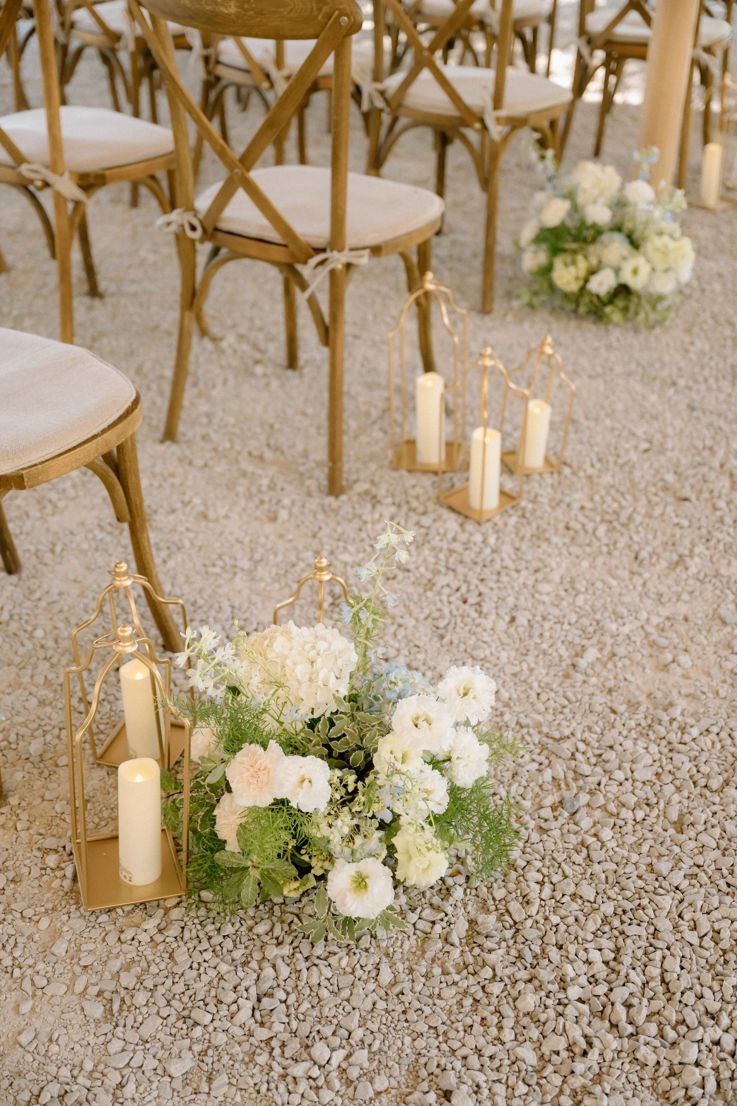 Assise de mariage avec des chaises en bois, un centre de fleurs blanches, roses et vertes, et des bougies dans des lanternes dorées, sur un sol en gravier.