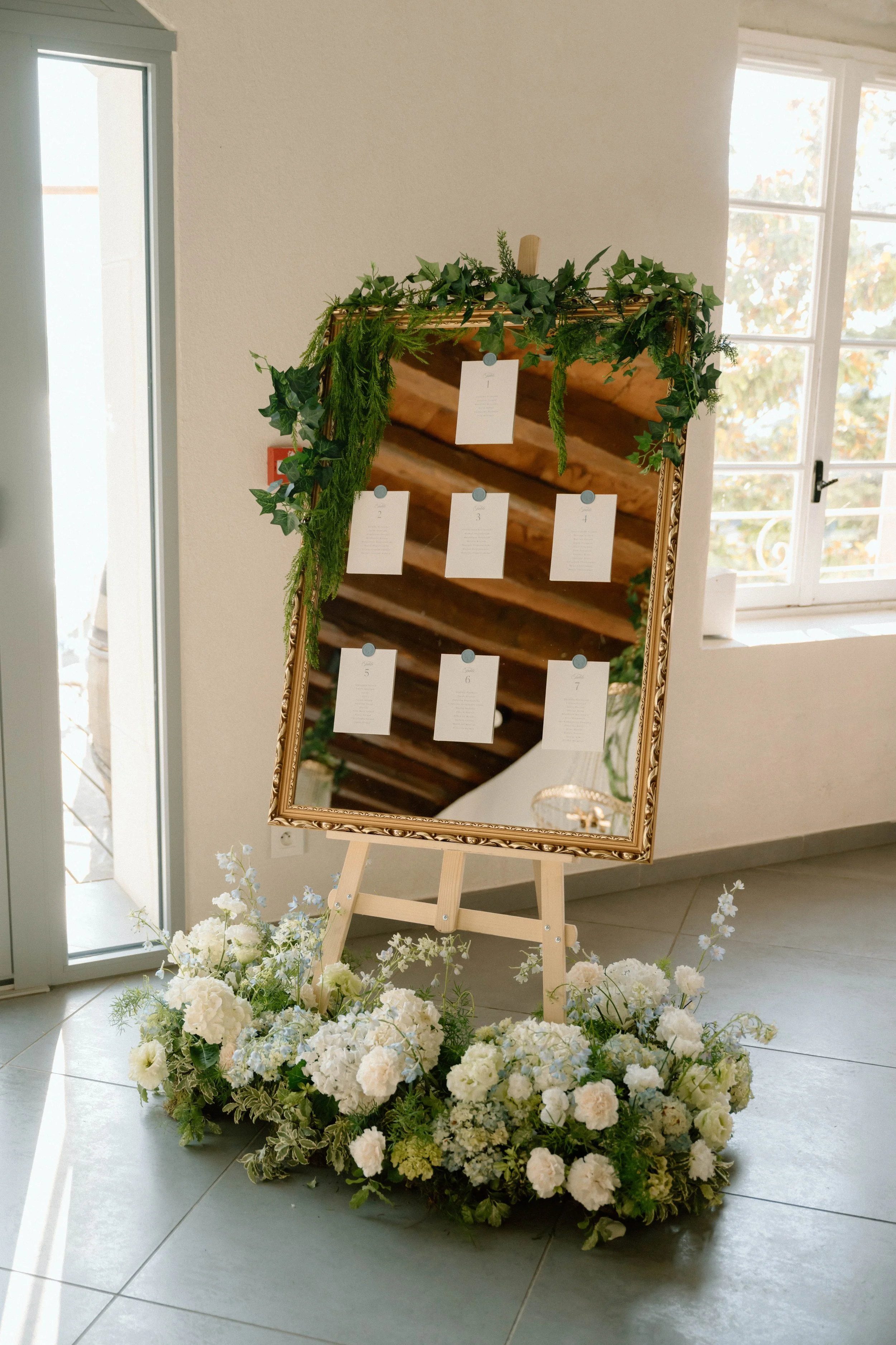 Un grand miroir décoré avec des fleurs blanches et des feuillages verts, placé sur un support en bois, dans une pièce lumineuse avec des fenêtres.