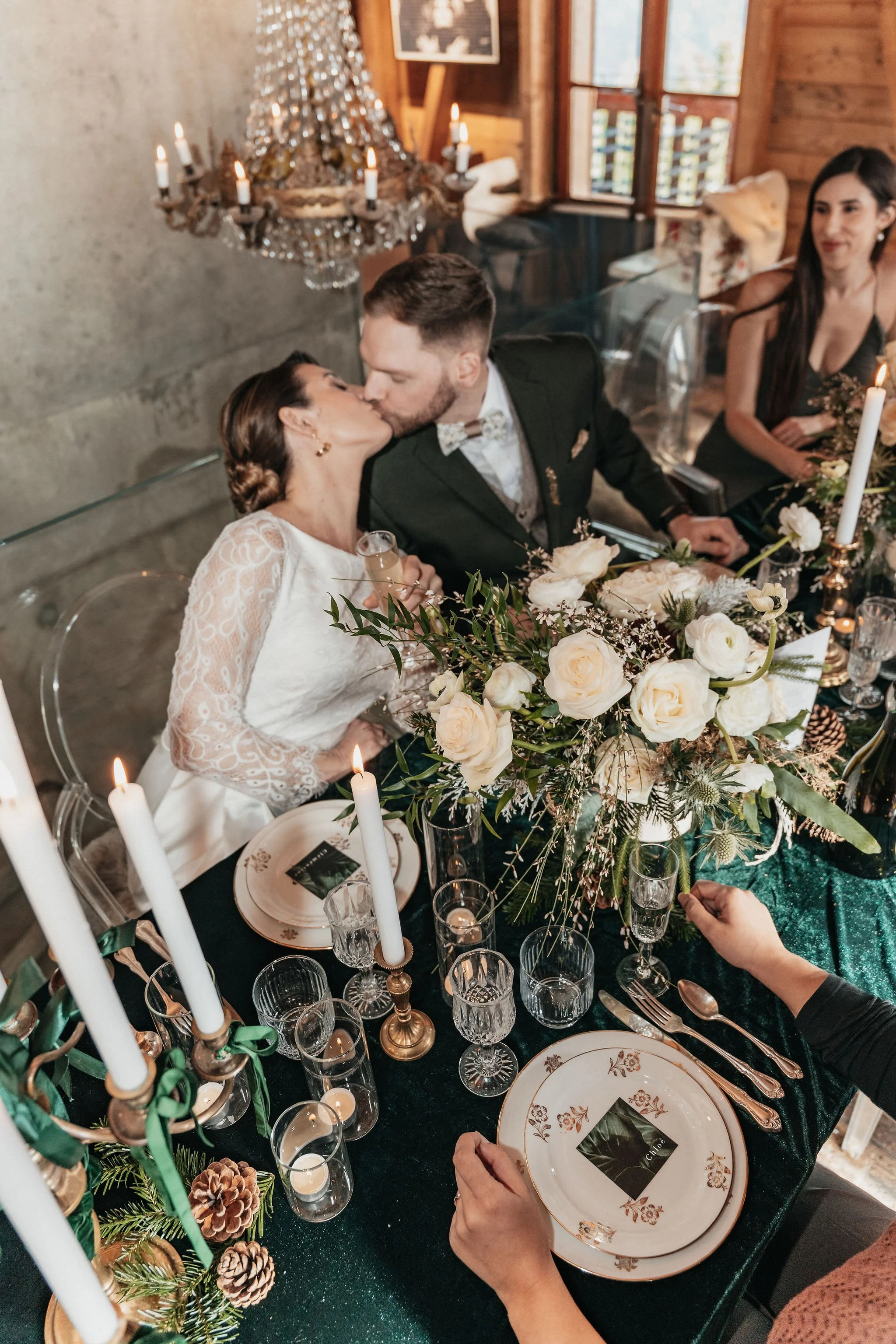 Un couple en habits de mariage s'embrasse lors d'une fête de mariage. La table est décorée avec des bougies, un bouquet de roses blanches, et de la vaisselle élégante.