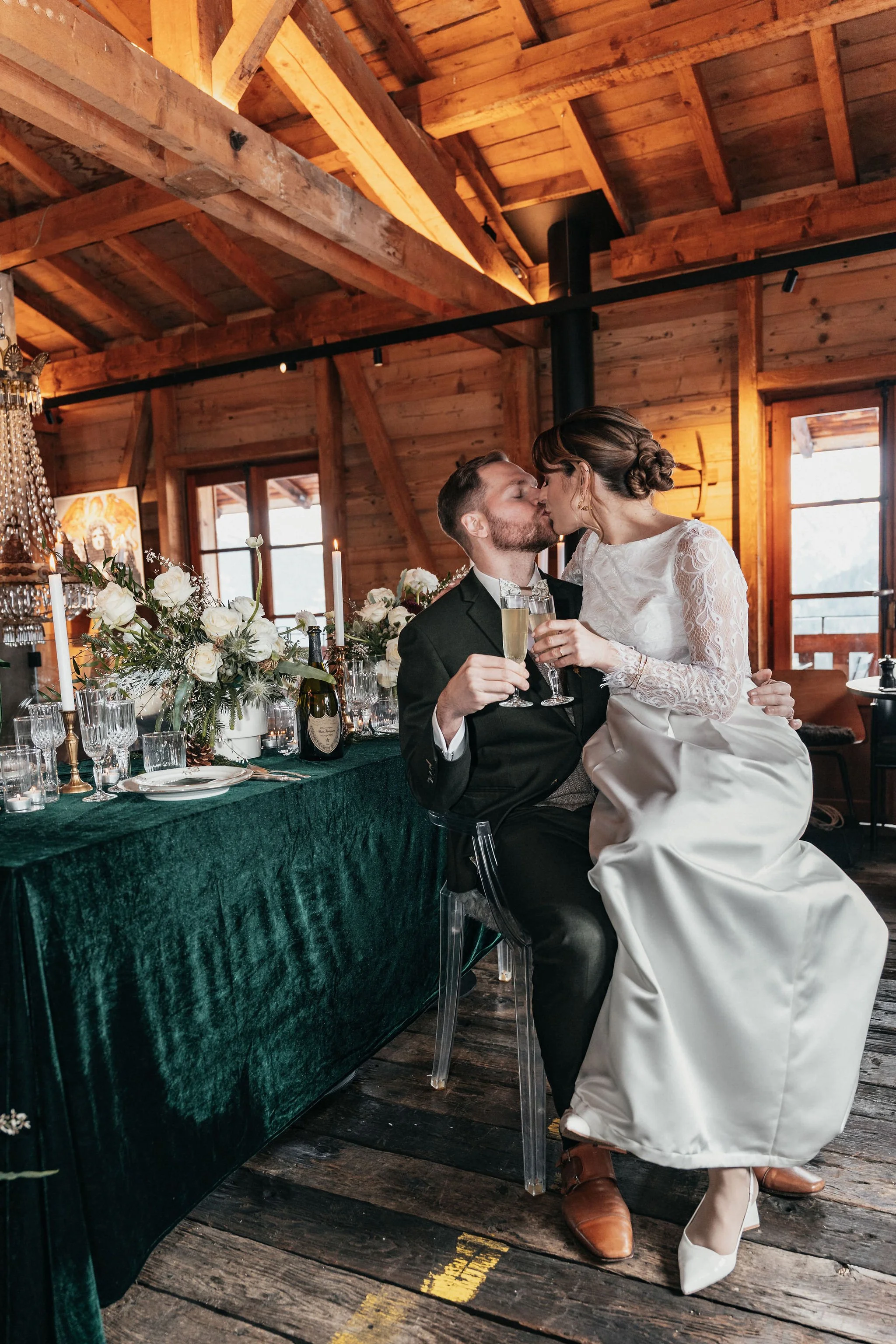 Un couple de mariés s'embrassant et trinquant lors de leur mariage dans une salle en bois chaleureuse, décorée de fleurs et de chandeliers.