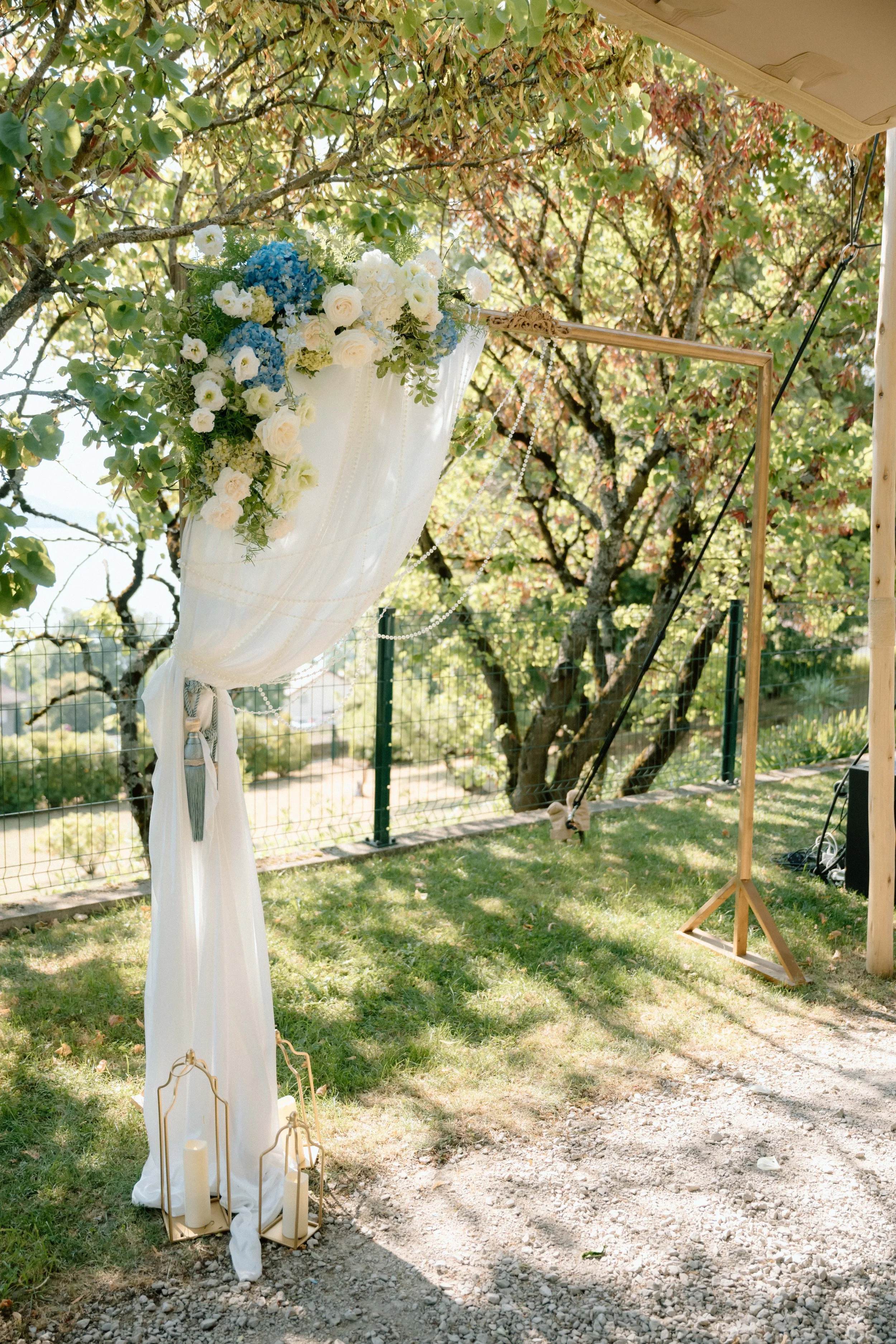 Décoration de mariage en plein air avec un arrangement floral blanc et bleu suspendu, un drap blanc avec des bougies dans des supports dorés, et un cadre en bois pour altar dans un jardin ensoleillé avec des arbres.