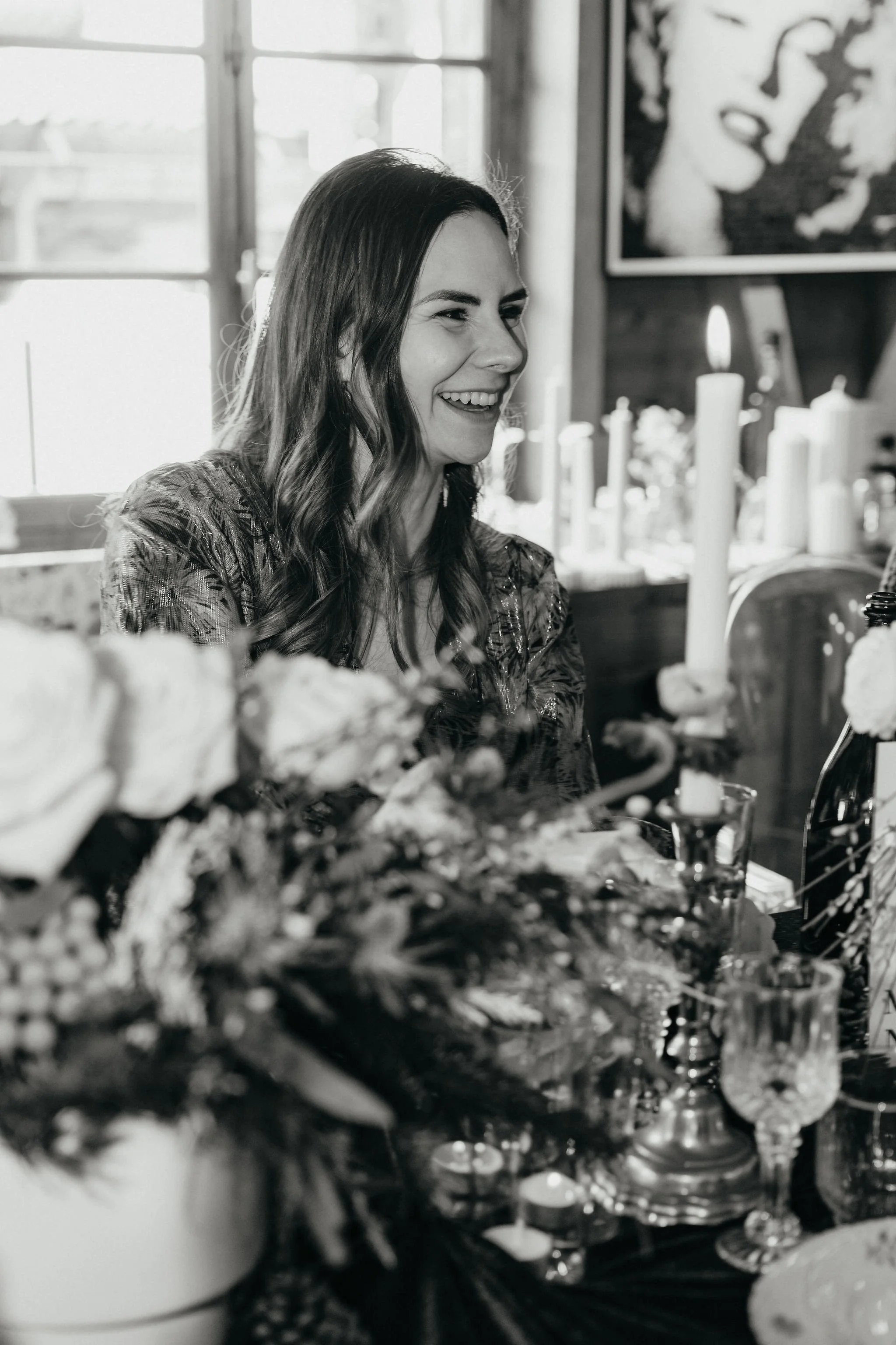 Une femme souriante assise parmi des décorations de fête, avec des bougies et des bouquets de fleurs sur la table, dans un intérieur lumineux.
