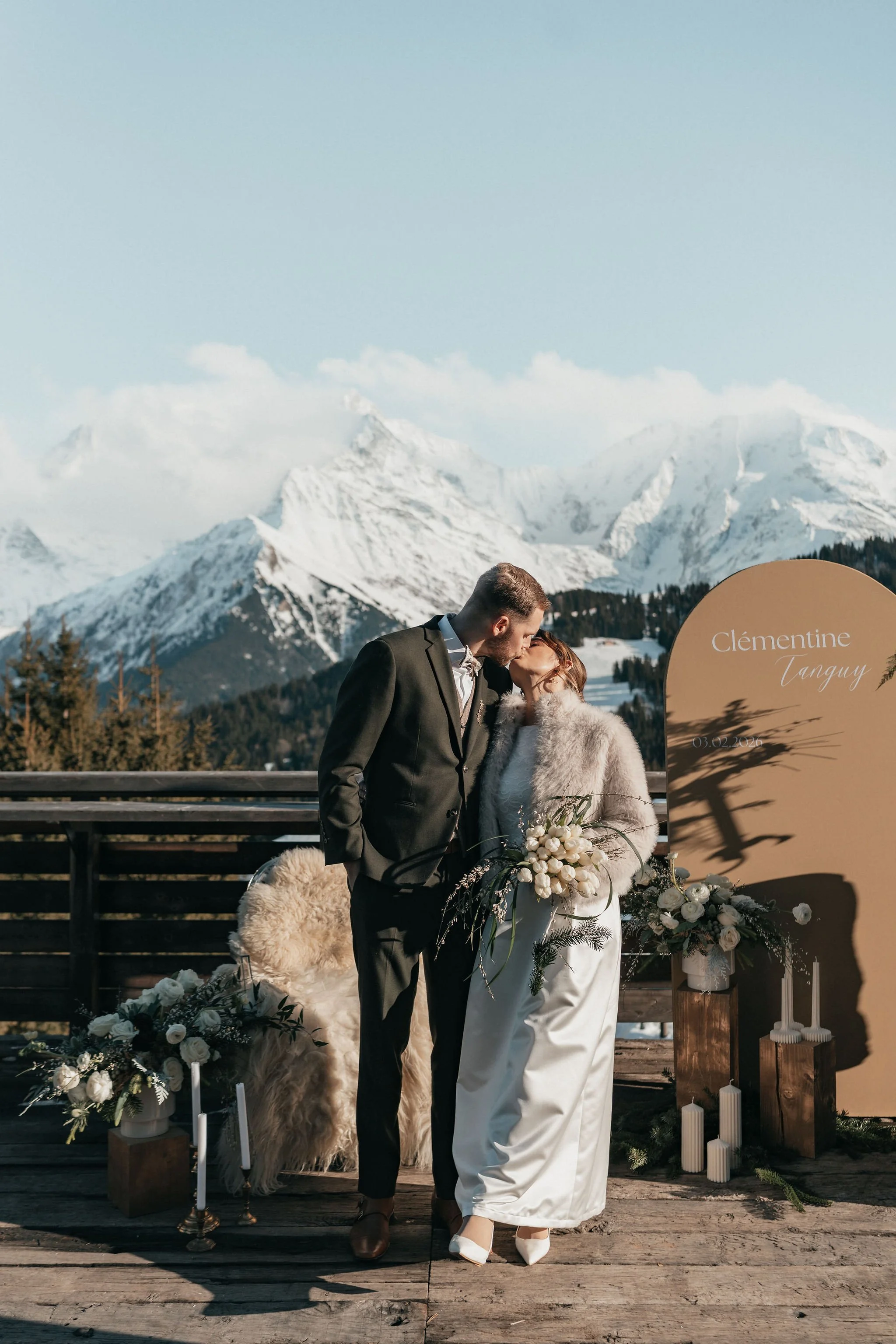 Un couple en vêtements de mariage s'embrasse devant un décor de mariage en montagne avec des sommets enneigés en arrière-plan.