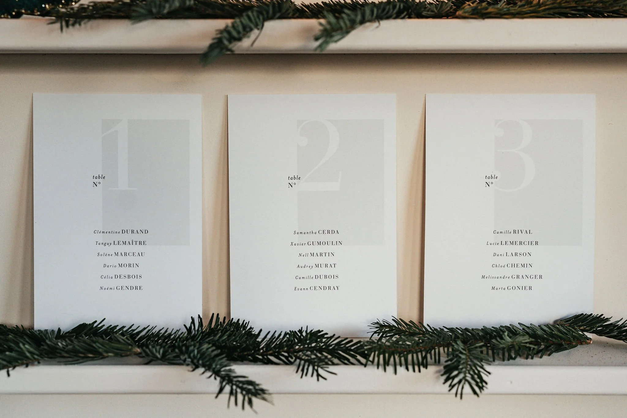 Trois affiches blanches avec des listes de noms et numéros de tables, décorées avec des branches de sapin dans un contexte festif.