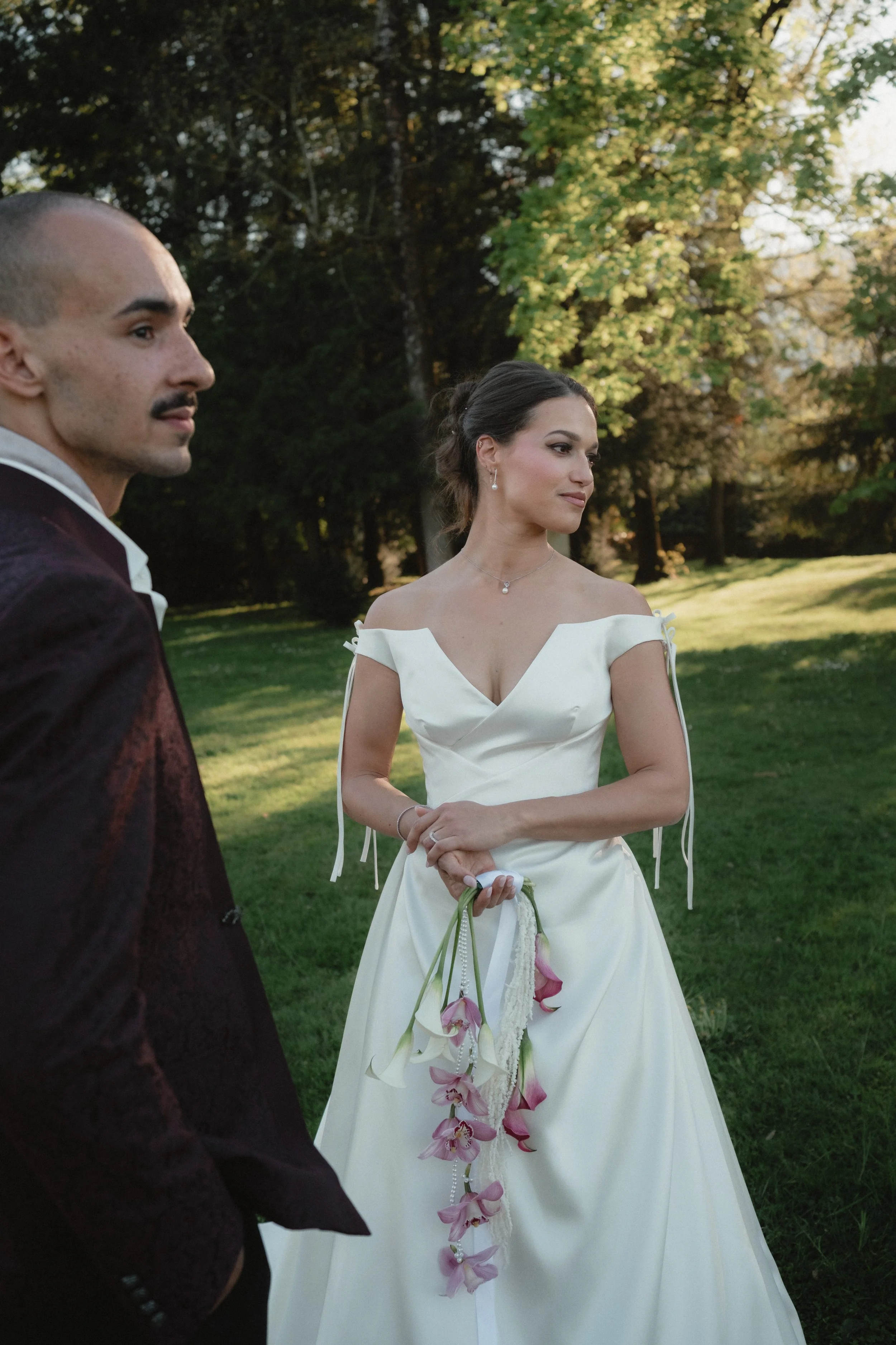 Un couple habillé élégamment, l'homme en costume sombre et la femme en robe de mariage blanche, se tenant dans un parc verdoyant avec des arbres aux feuilles vertes et jaunes.