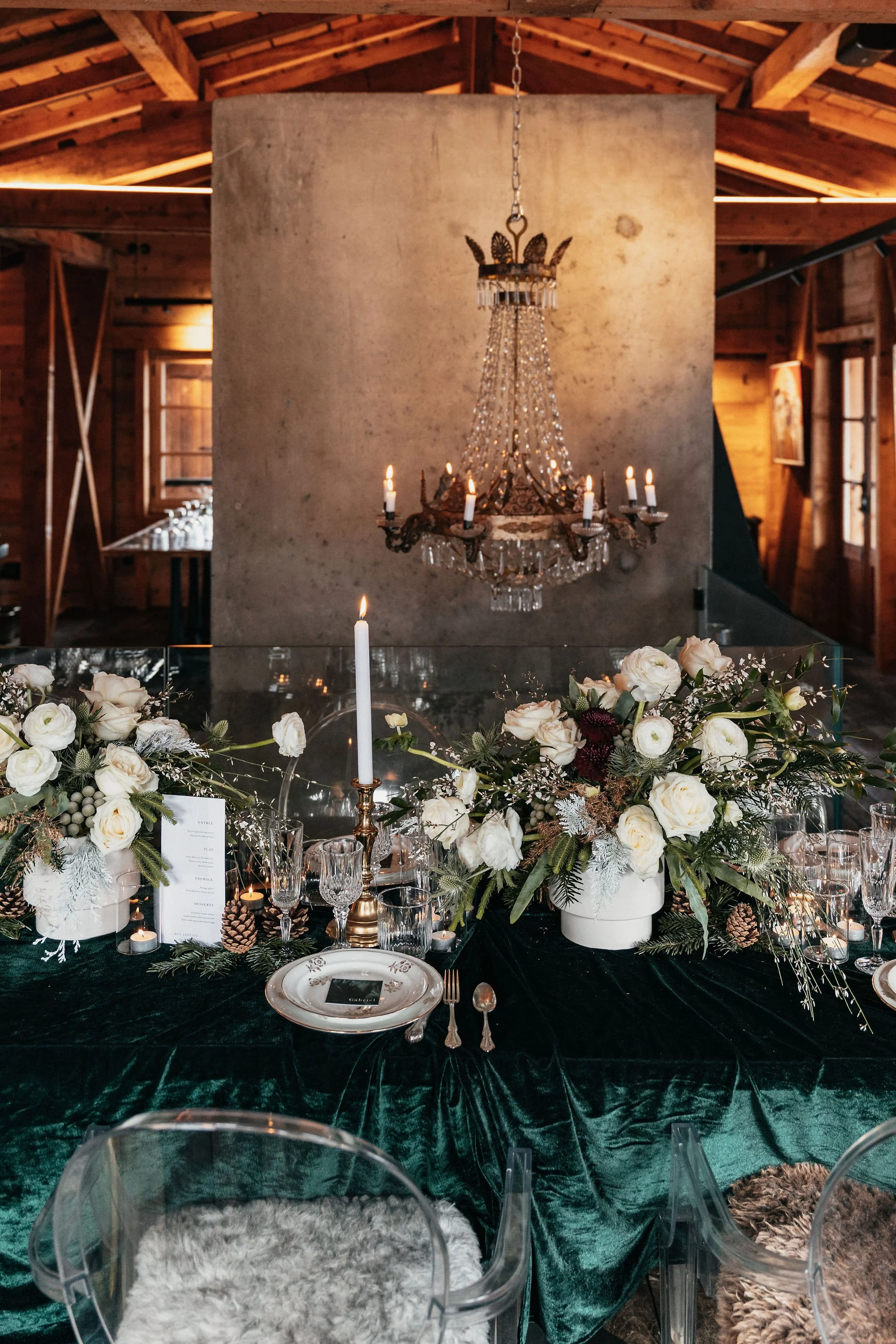 Une salle de mariage ou de réception avec une table décorée de fleurs blanches, bougies et vaisselle élégante, un chandelier suspendu en cristal et un fond en bois avec une structure en chaume au plafond.