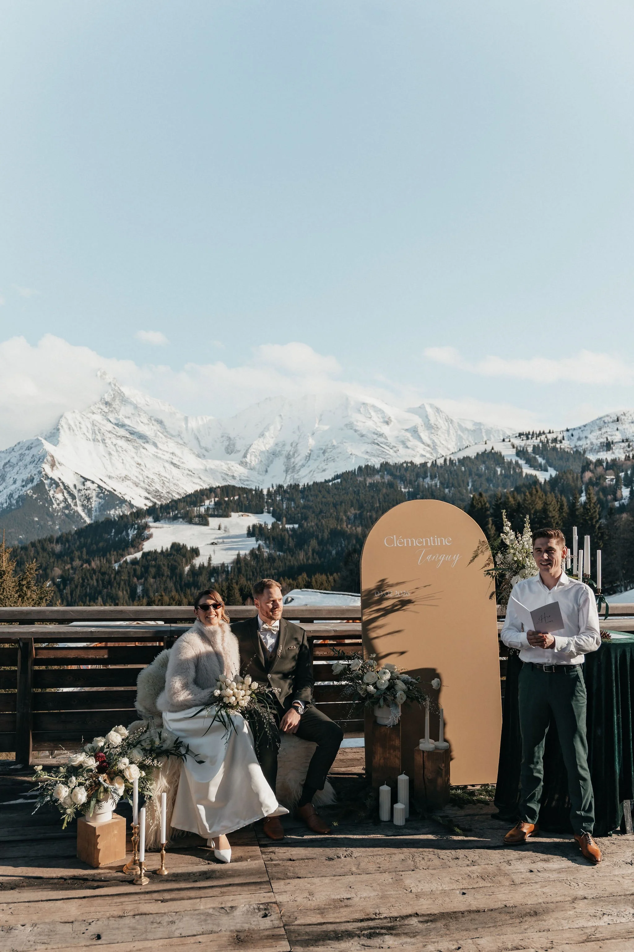 Un couple en costume de mariage assis à une cérémonie en plein air dans un paysage de montagnes enneigées, avec un officiant à côté et des décorations florales et bougies autour.