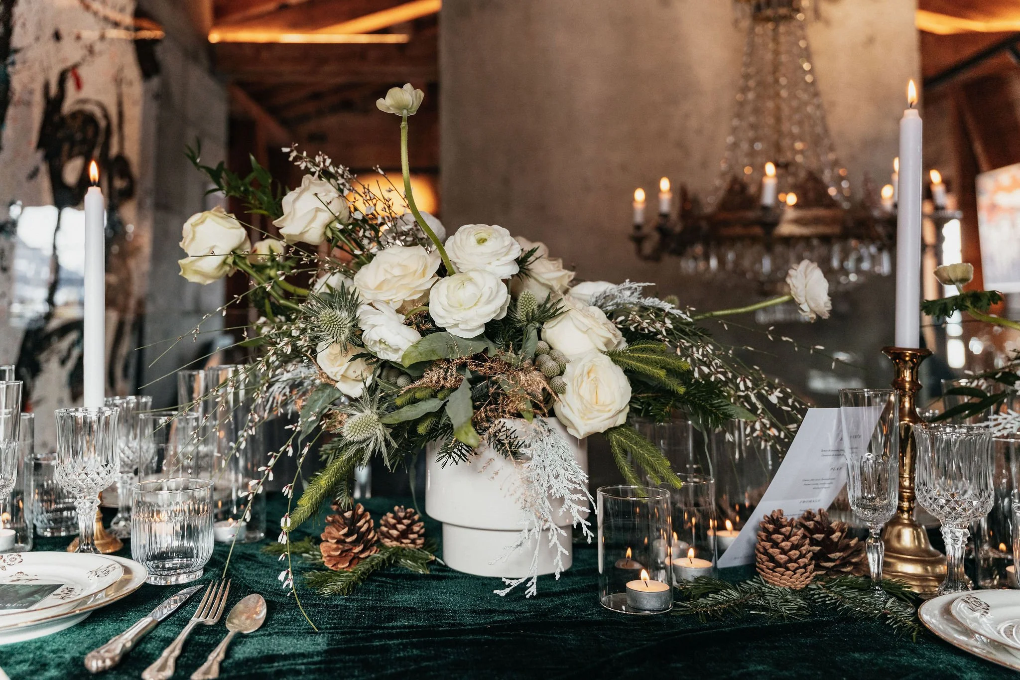 Centre de table avec des fleurs blanches, bougies, bougeoirs, pommes de pin sur une nappe verte lors d'un dîner.
