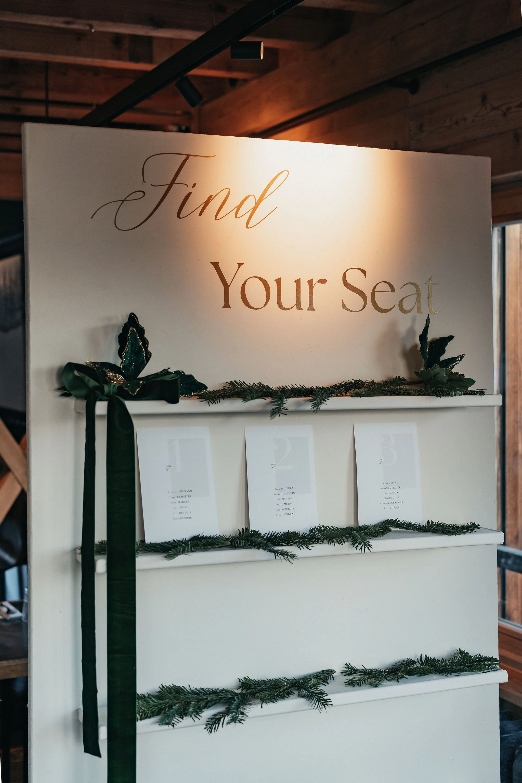 Une affiche de mariage avec l'inscription "Find Your Seat" décorée de branches de sapin et un ruban vert, indiquant un système de seating pour un événement chic et festif.