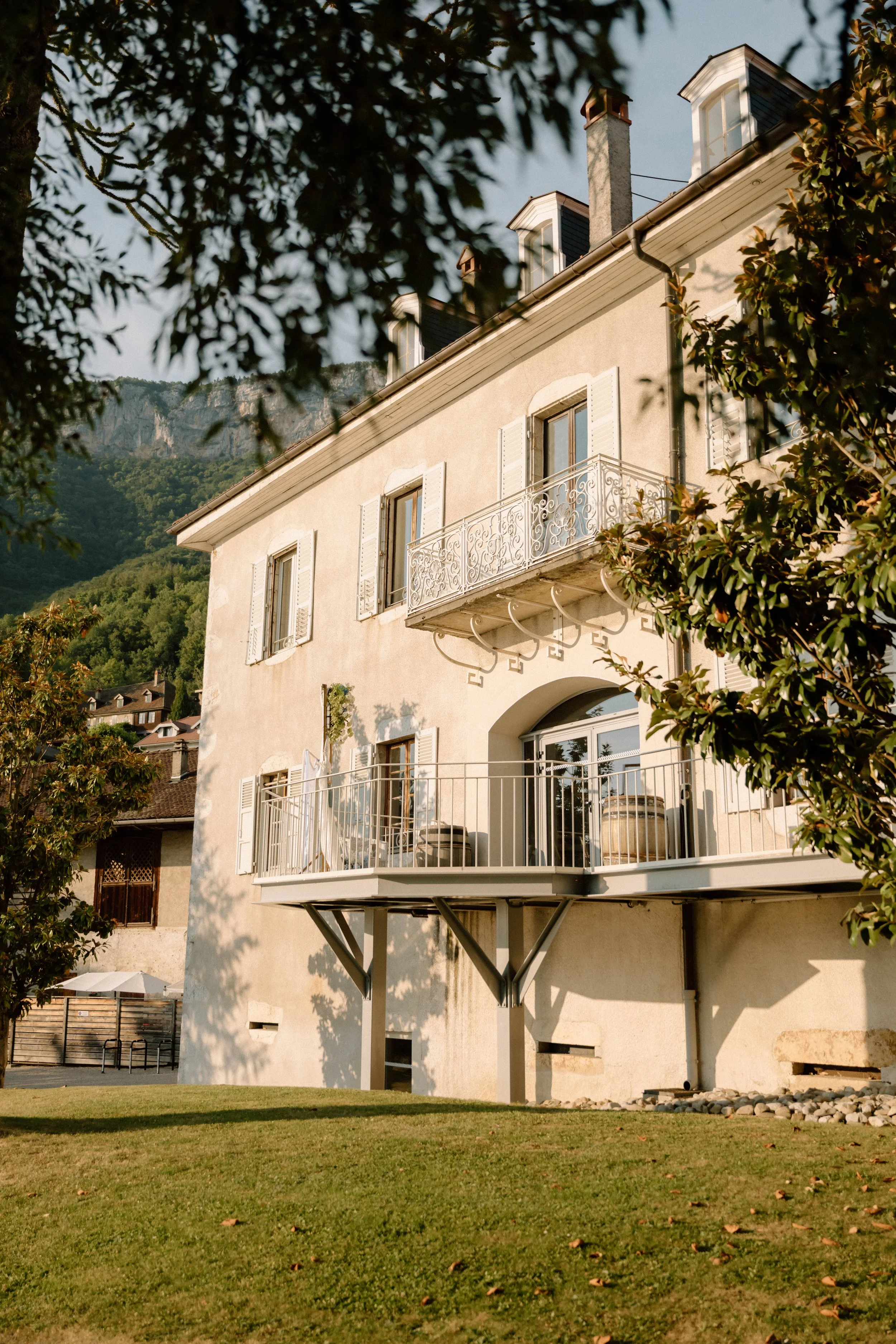 Immeuble résidentiel de style européen avec plusieurs fenêtres à volets blancs, un balcon en fer forgé, entouré d'arbres et de montagnes en arrière-plan, sous un ciel en fin d'après-midi.