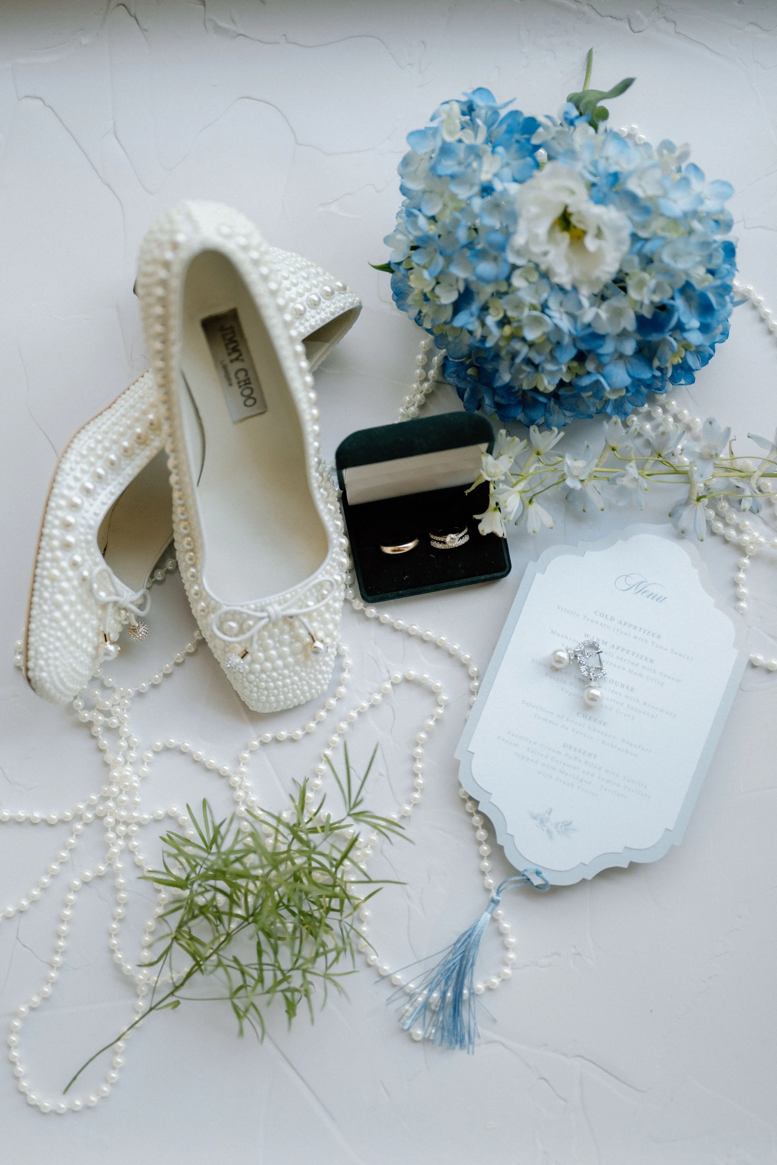 Chaussures de mariage blanches décorées de perles, bouquet de fleurs bleues et blanches, registres de mariage, perles, carte de menu, bagues dans une boîte noire, décoration florale et pendentif.