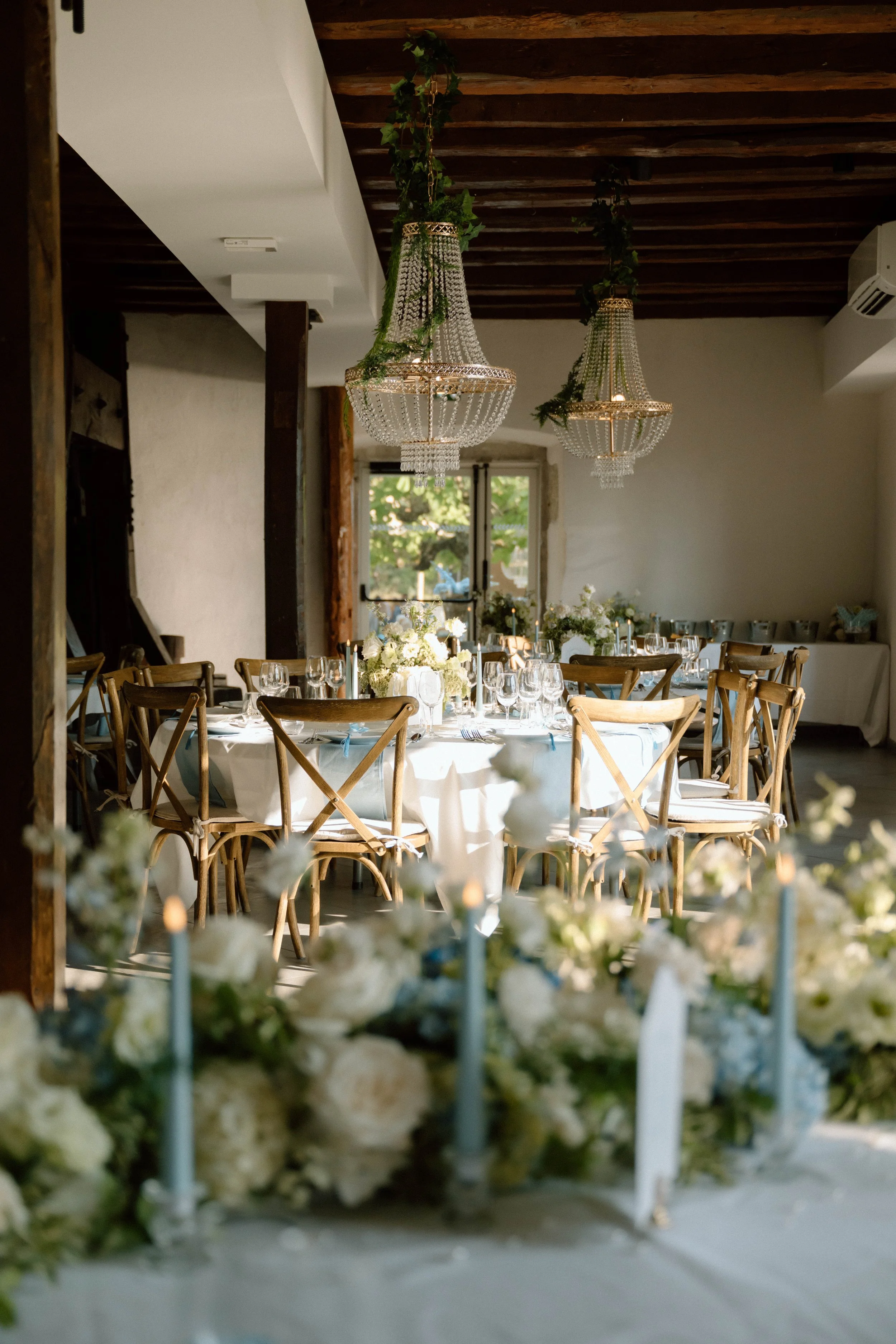 Salle de réception décorée pour un événement, avec table dressée, chandeliers en cristal suspendus, et fleurs blanches au centre, dans une pièce lumineuse avec grande fenêtre.