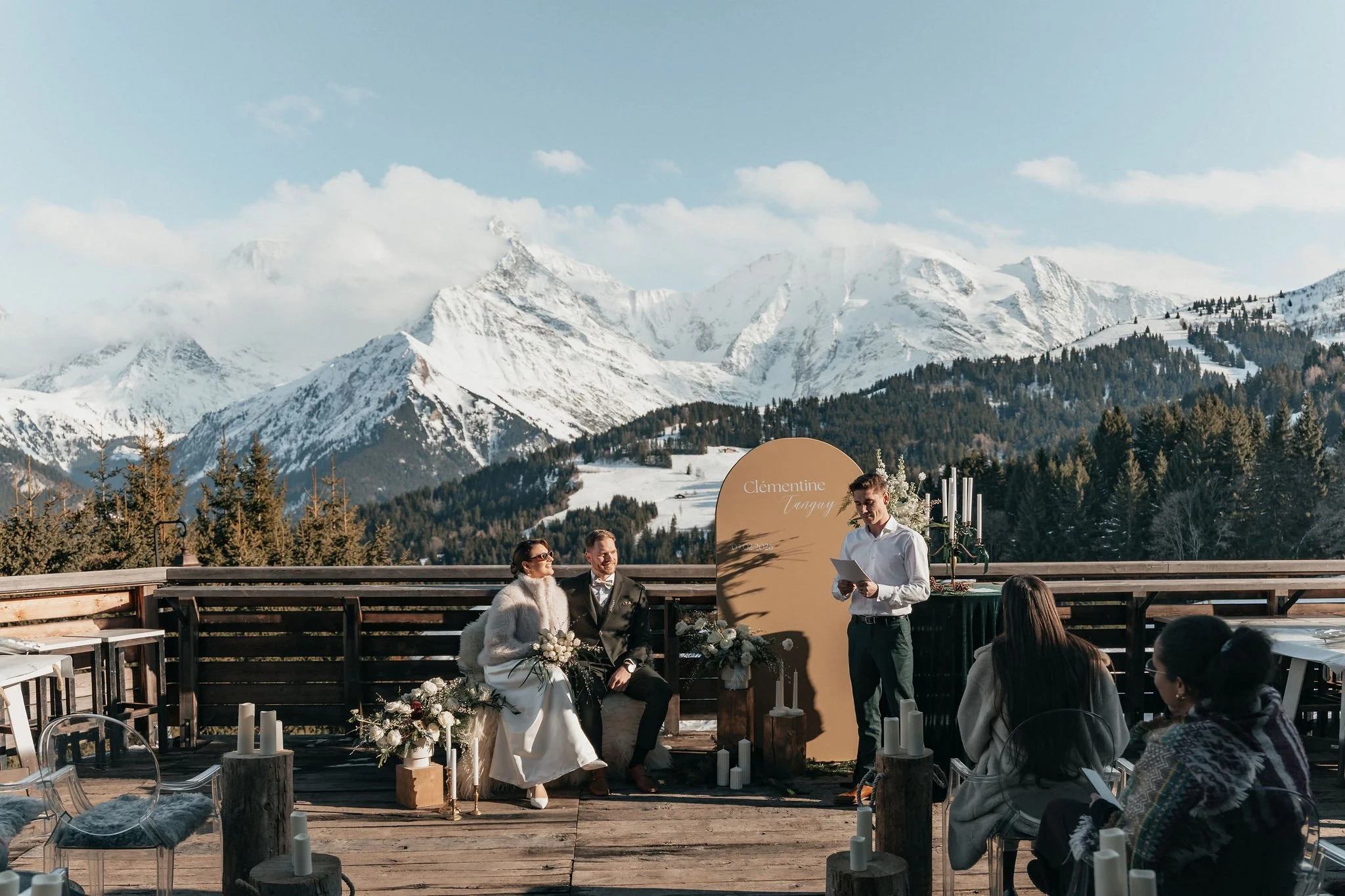 Cérémonie de mariage en plein air avec montagnes enneigées en arrière-plan, couple assis lors de la cérémonie, officiant lisant un discours, invités assis, décor nature avec bougies et fleurs.