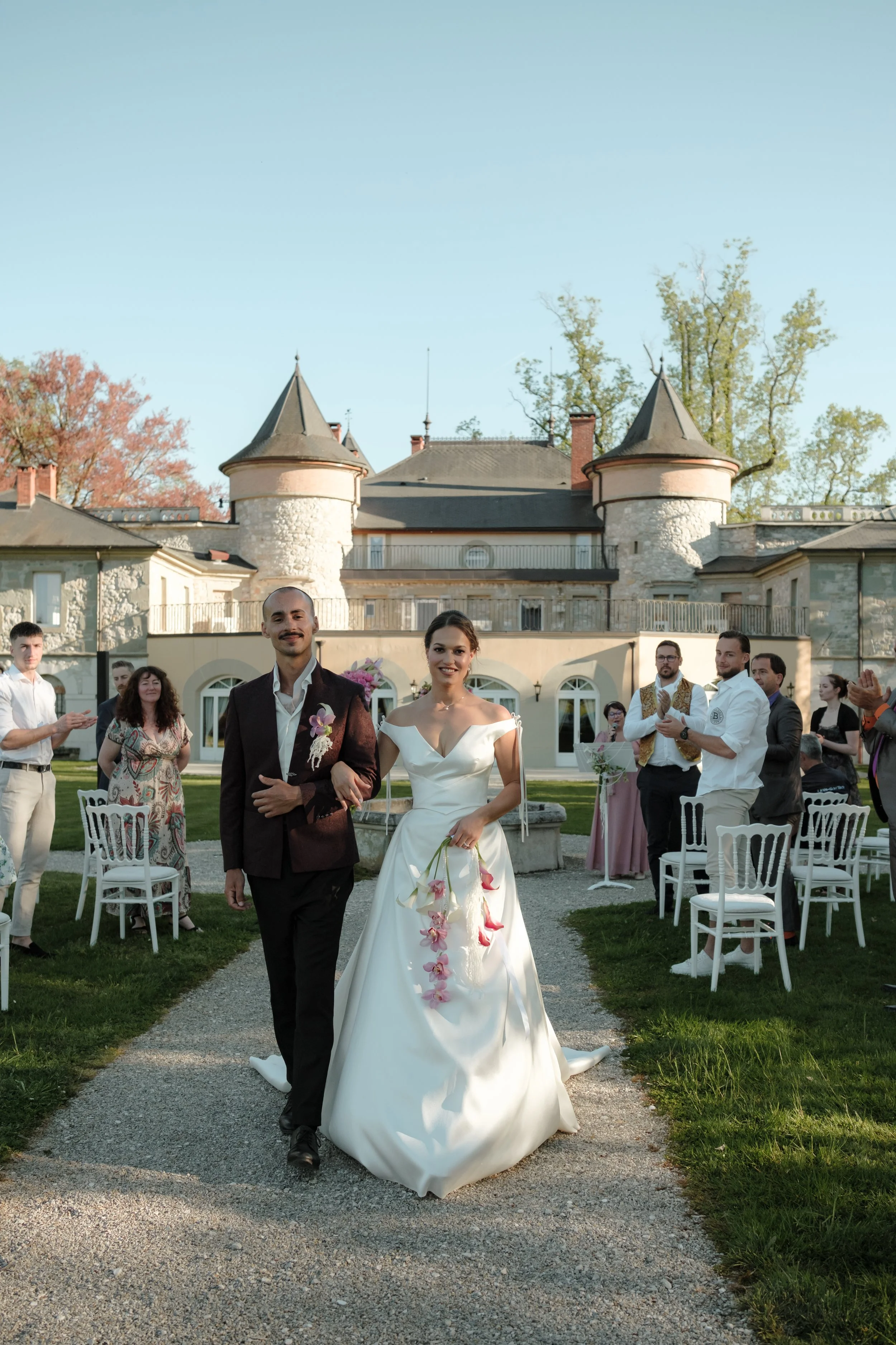 Un couple de mariés marche main dans la main sur un chemin lors d'un mariage en extérieur, avec un château en pierre en arrière-plan et des invités qui applaudissent autour. La femme porte une robe de mariée blanche et le homme un costume noir.
