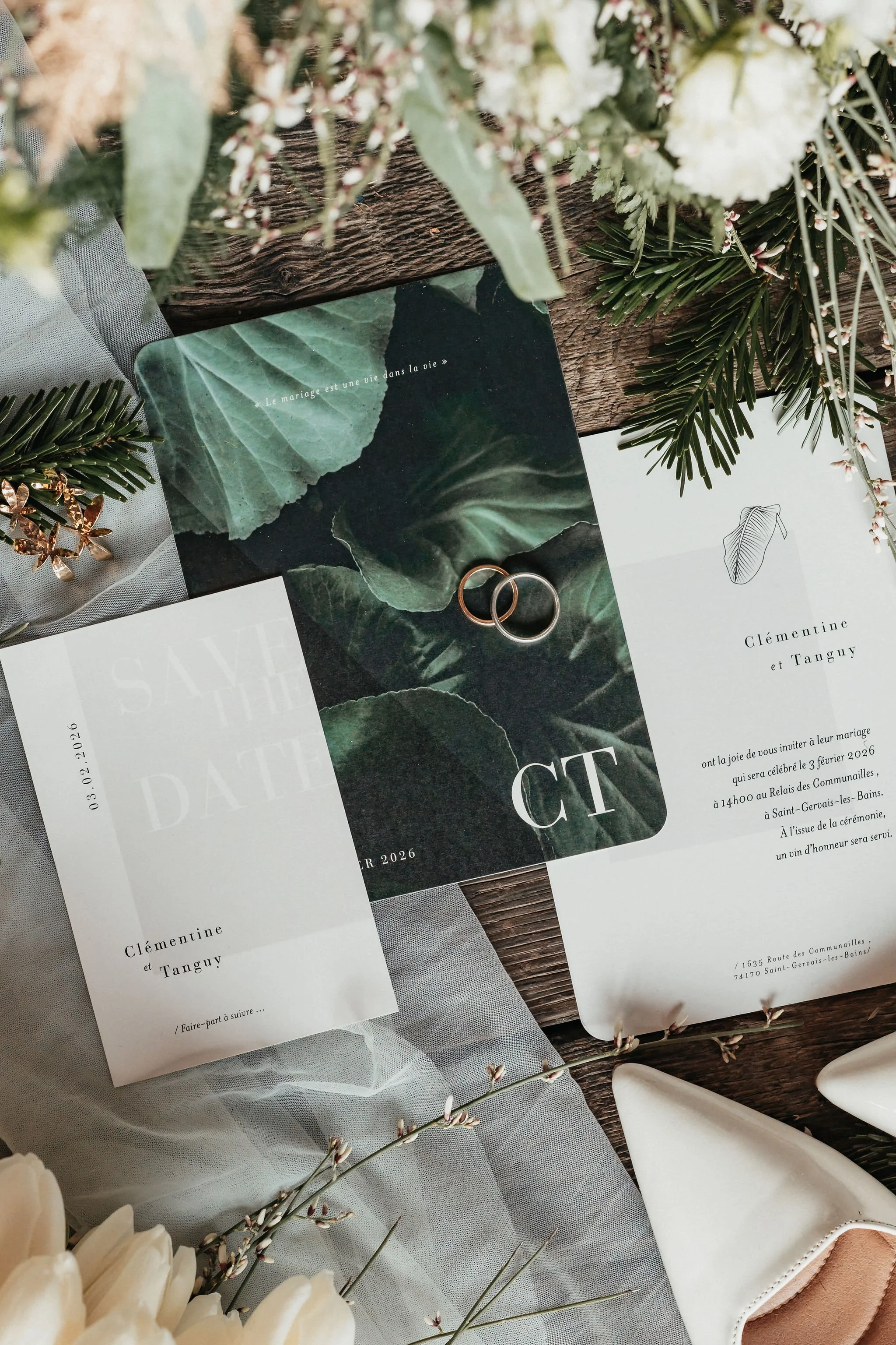 Invitations de mariage avec des anneaux, fleurs blanches et feuillage vert, posées sur une surface en bois.