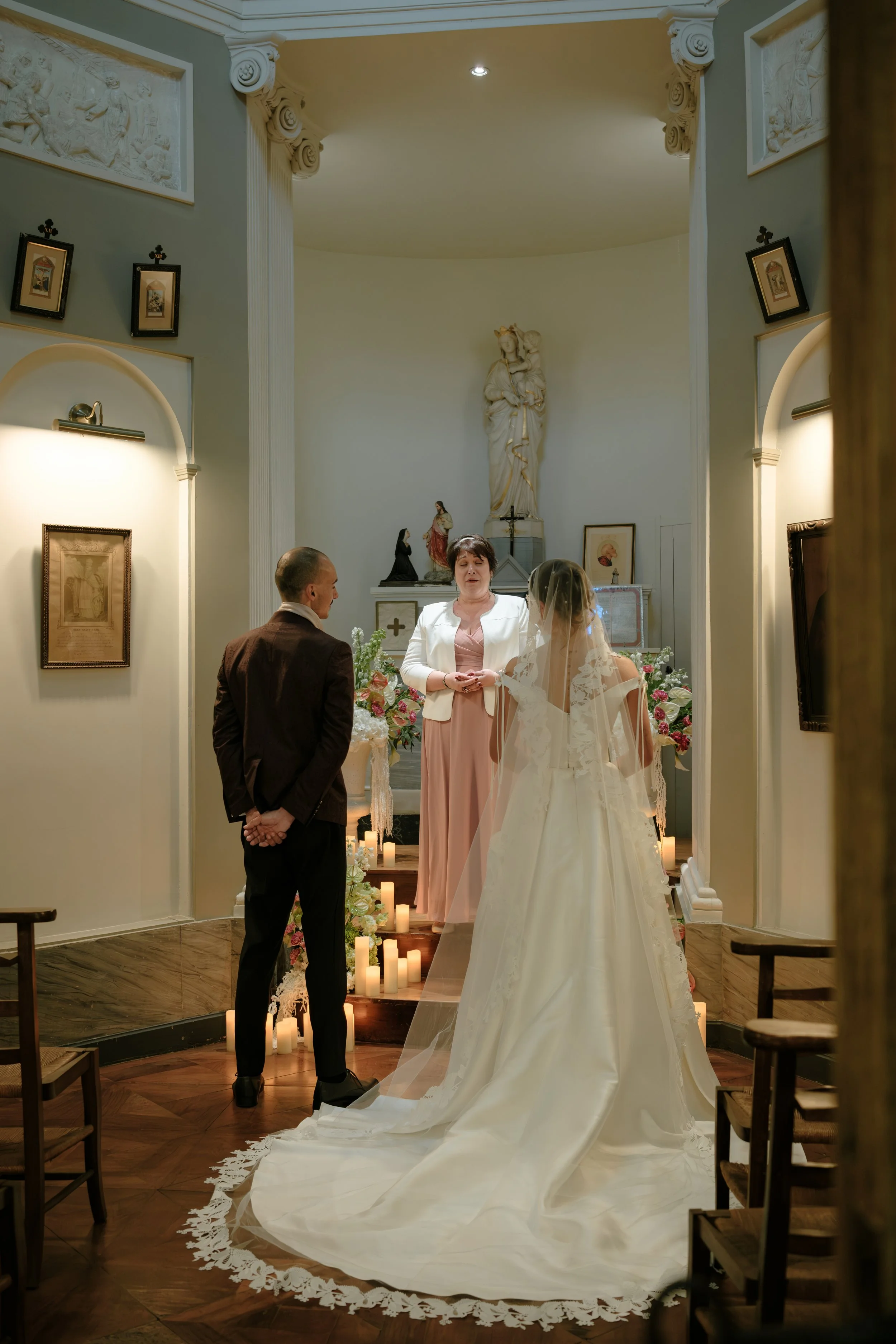 Cérémonie de mariage dans une église, avec un couple de mariés, une officiante et un témoin, devant un autel orné de statues religieuses et de bougies.
