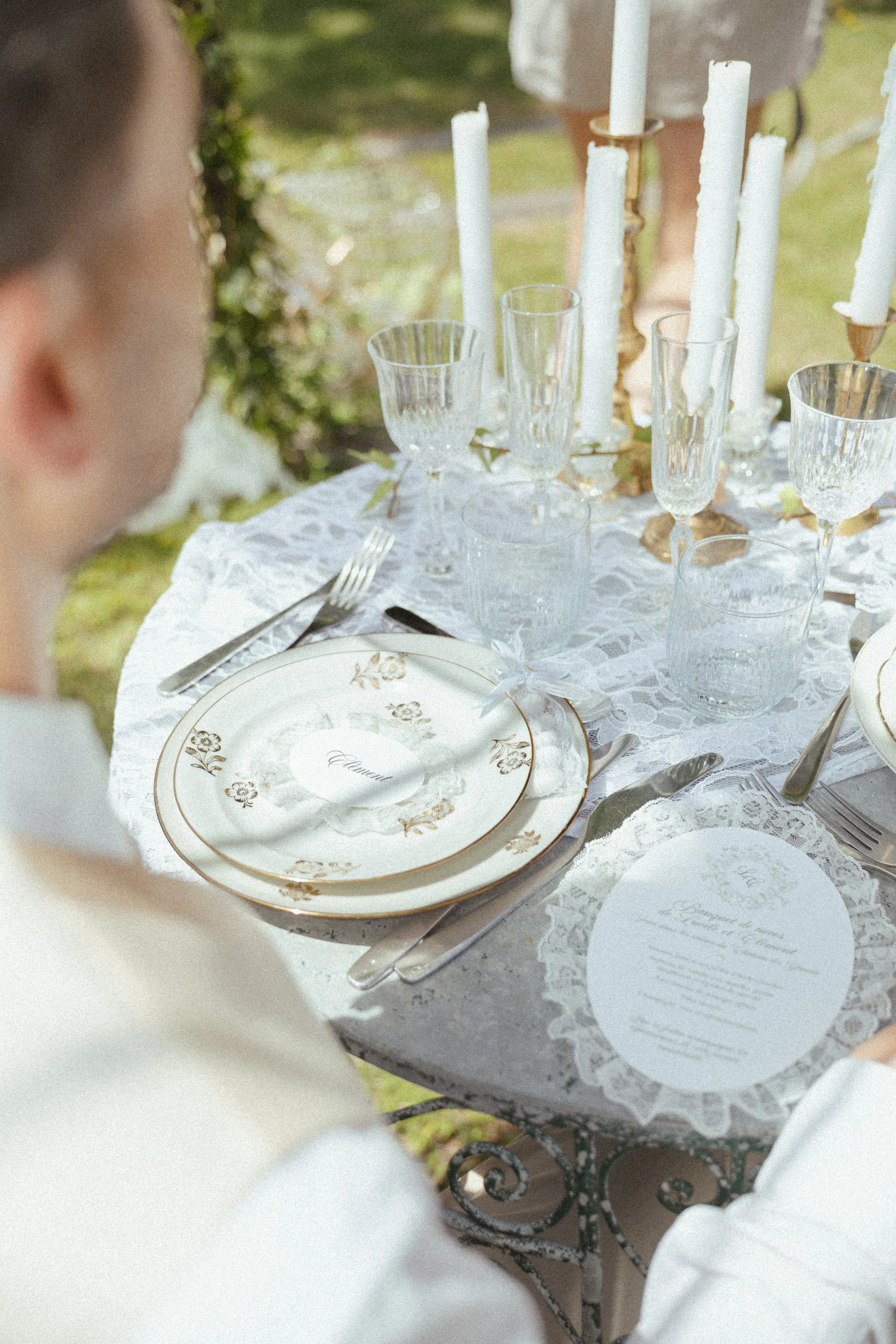 Une table élégante dressée pour un repas en plein air avec de la vaisselle en porcelaine blanche ornée de motifs dorés, des verres en cristal, des couverts en argent, des bougies blanches dans des chandeliers en or, et un menu ou un nom de plat écrit