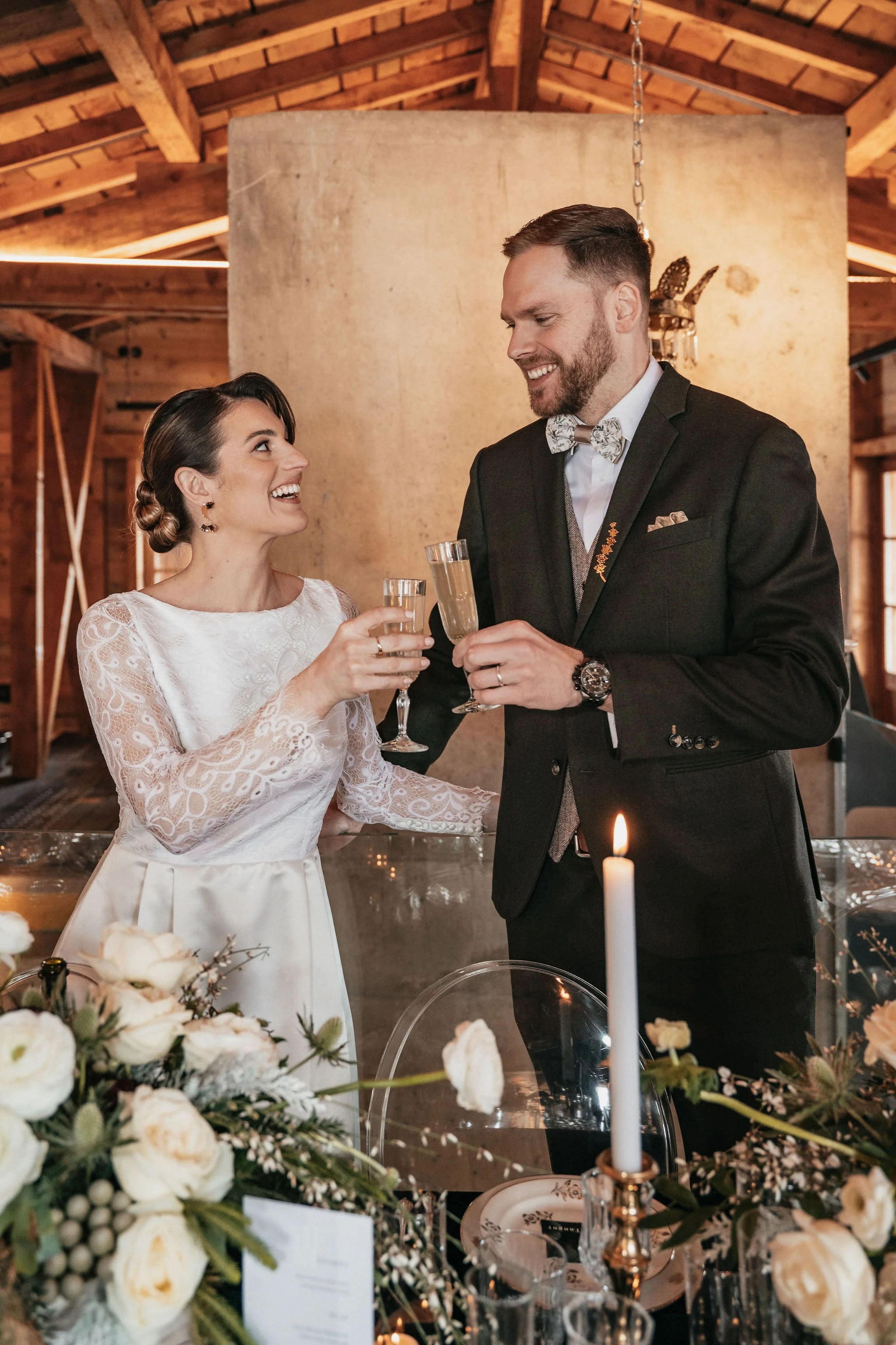 Un couple de mariage, souriant et levant leurs flûtes, en train de faire un toast dans une ambiance chaleureuse et élégante avec des fleurs blanches et une bougie allumée sur la table.