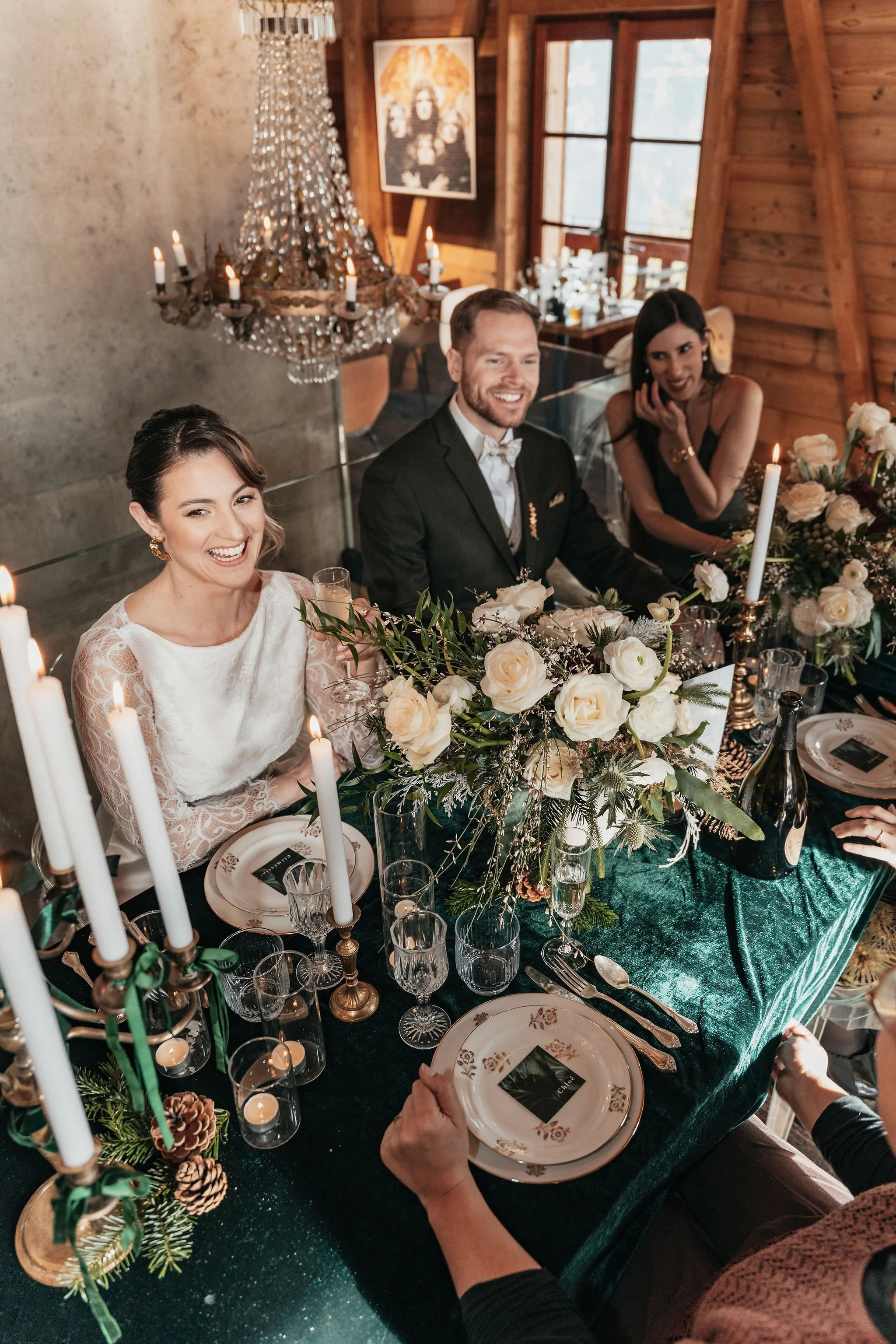 Groupe de personnes à une table décorée pour un repas, avec des fleurs blanches, chandelles et vaisselle élégante, dans une maison en bois. Deux femmes et un homme sourient et discutent dans une ambiance chaleureuse.