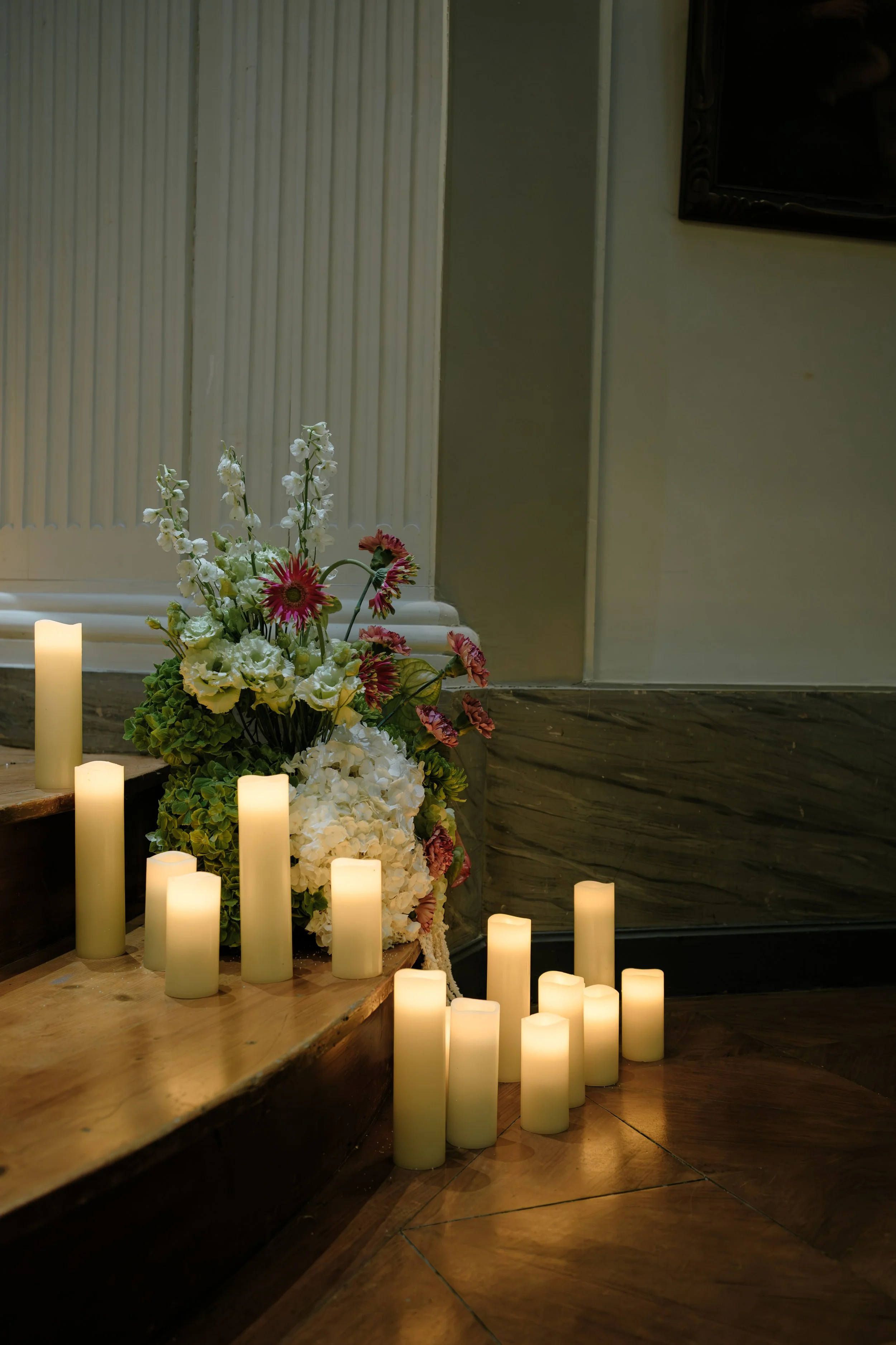 Un arrangement floral avec des bougies allumées autour, placée sur une table en bois, dans un intérieur, avec un mur blanc derrière.
