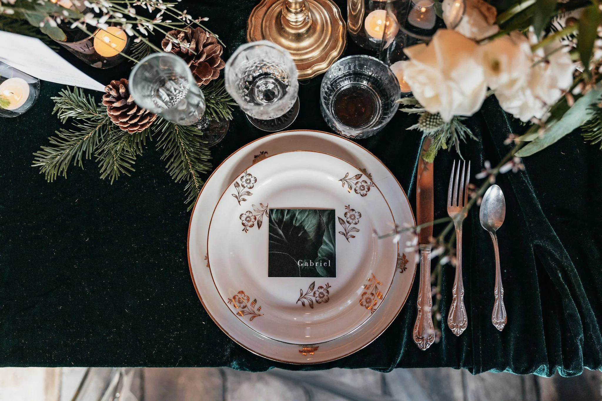 Table élégamment dressée avec une assiette en porcelaine blanche à motif floral doré, un verre d'eau, un verre à vin et des bougies, décorée de branches de sapin, pommes de pin et fleurs blanches, avec un fond en tissu vert foncé.