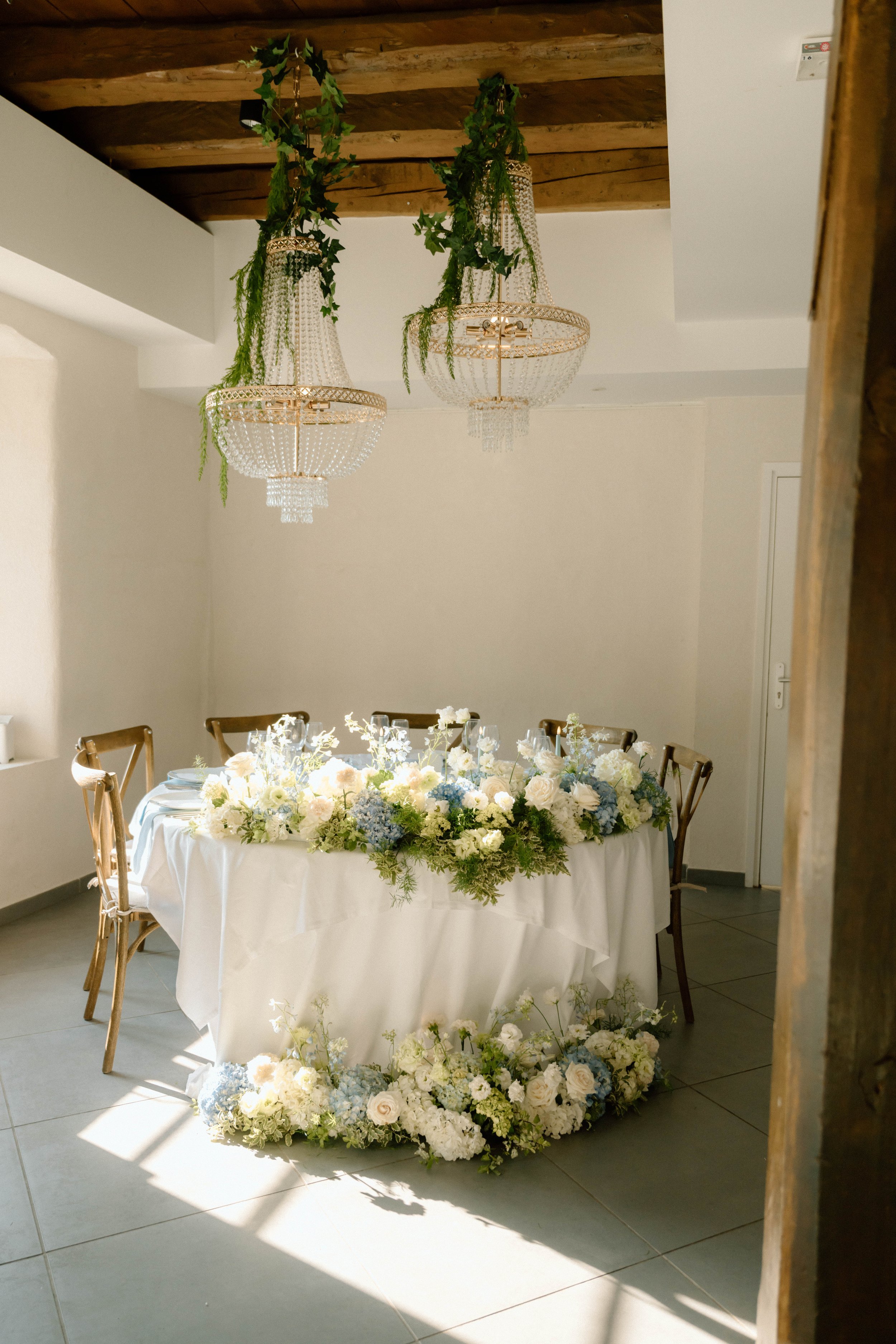 Une table de mariage décorée avec un centre de table rempli de fleurs blanches, bleues et vertes, avec deux lustres en cristal suspendus au plafond en bois. La pièce a des murs blancs et un sol en carreaux gris.