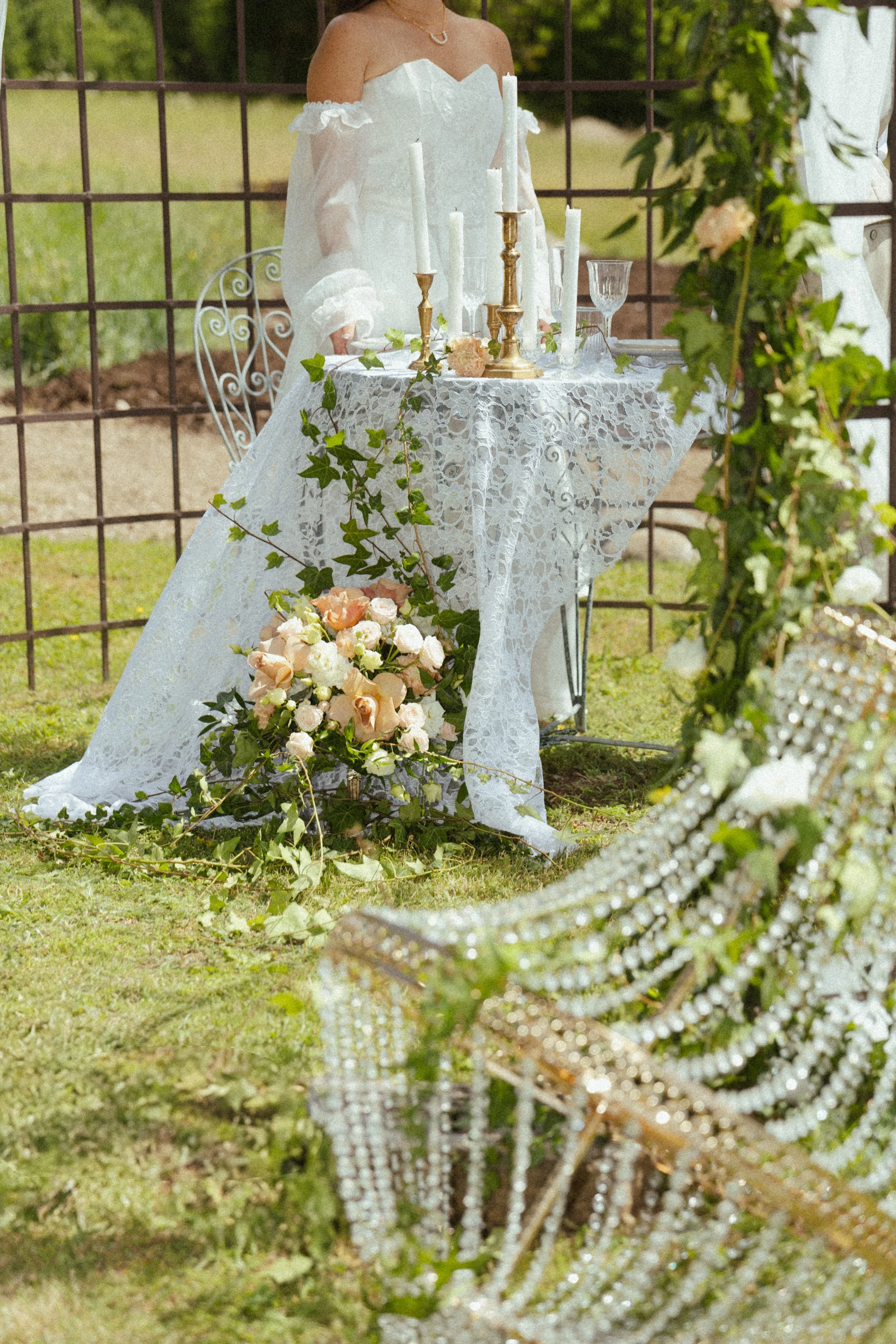 Une scène lors d'une cérémonie en plein air avec une table décorée de bougies, de fleurs et de voilage blanc, entourée de chaises en cristal et de verdure, en arrière-plan un cadre en métal et des arbres.