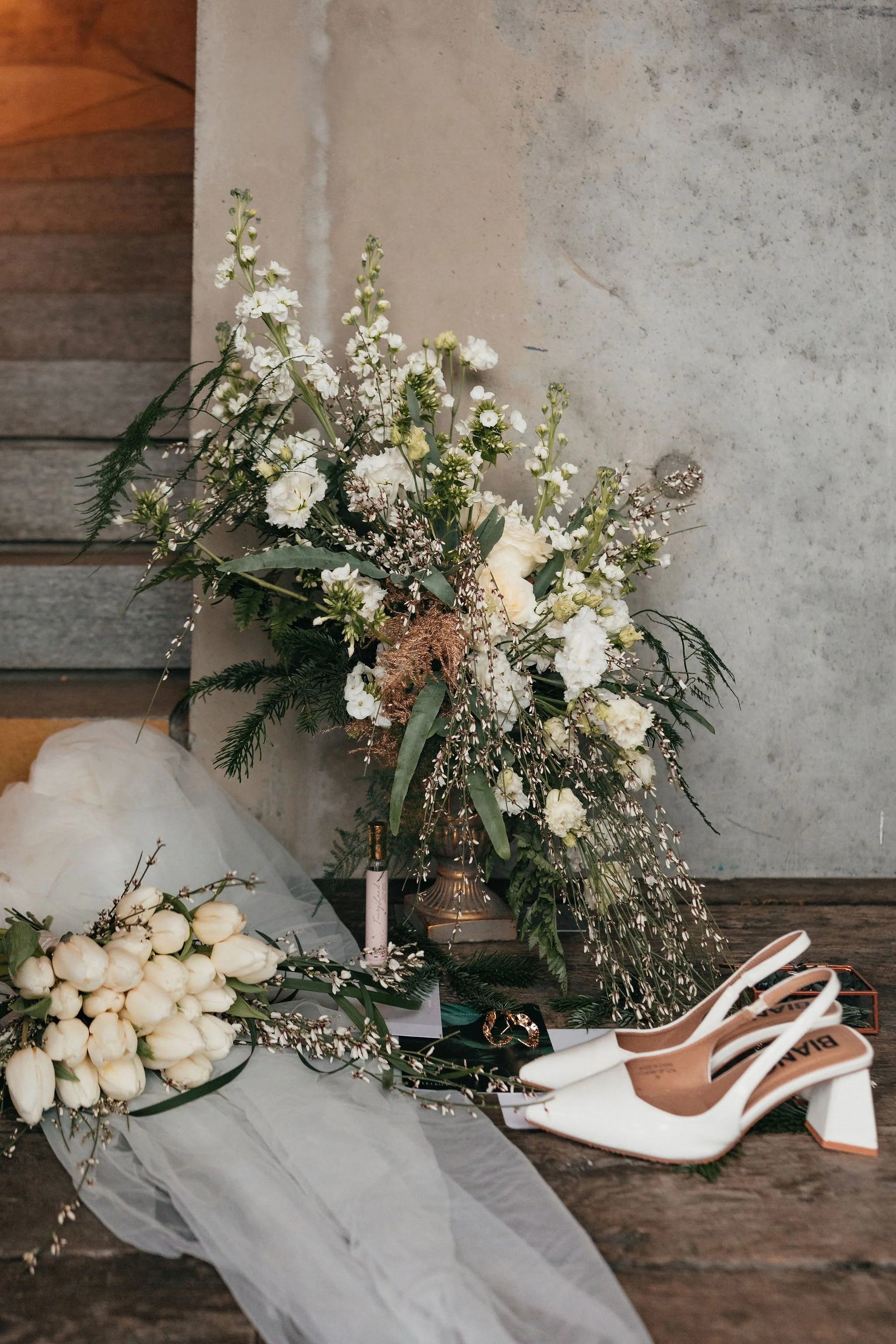 Bouquet de fleurs blanches, paire de chaussures blanches à talons, et un voile blanc posés sur un sol en bois.