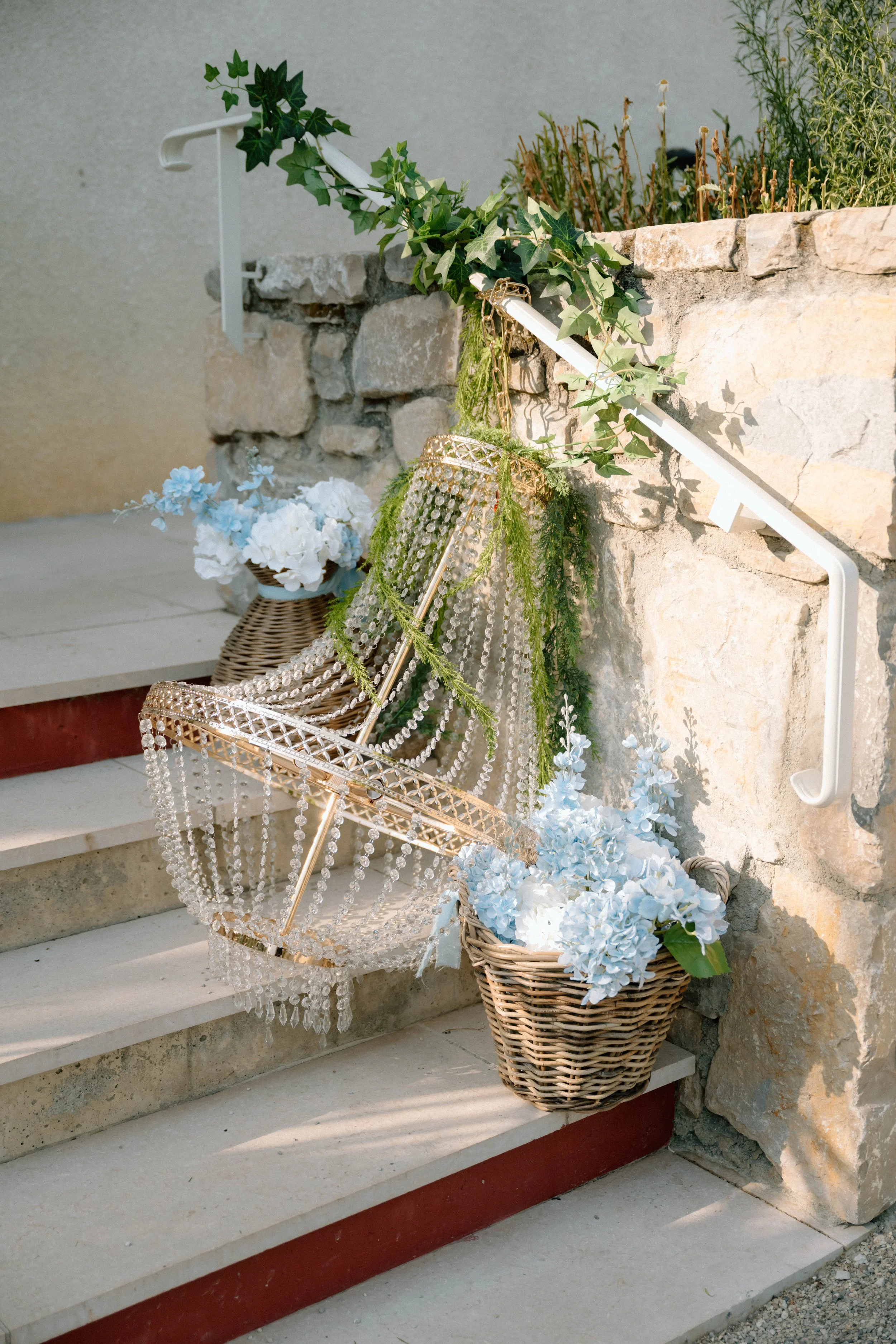 Escalier en pierre décoré de fleurs blanches dans un panier en osier, avec un chandelier en métal orné de guirlandes de perles et de plantes vertes, contre un mur en pierre.