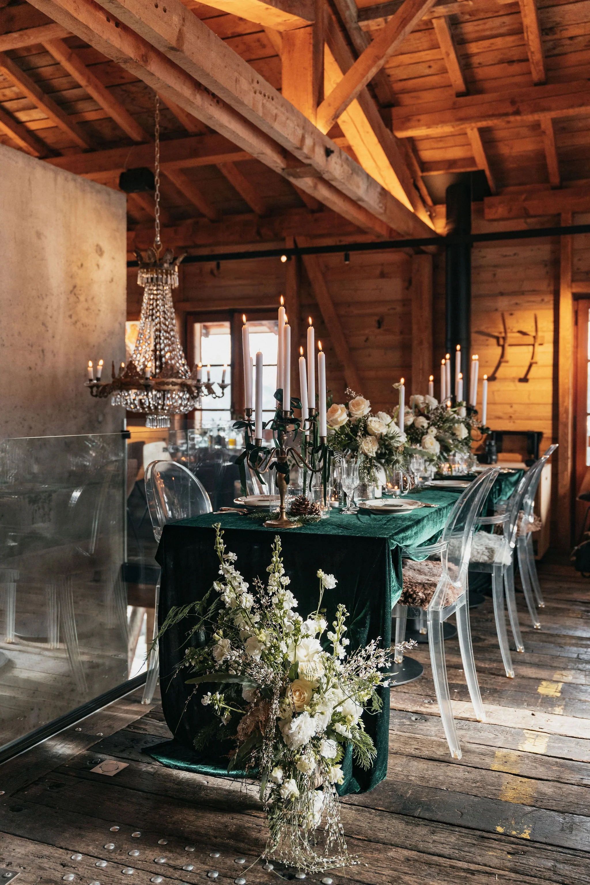 Une table décorée pour un dîner ou un événement, avec des chandeliers à bougies, des fleurs blanches et un filet dans un intérieur en bois avec un chandelier en cristal.