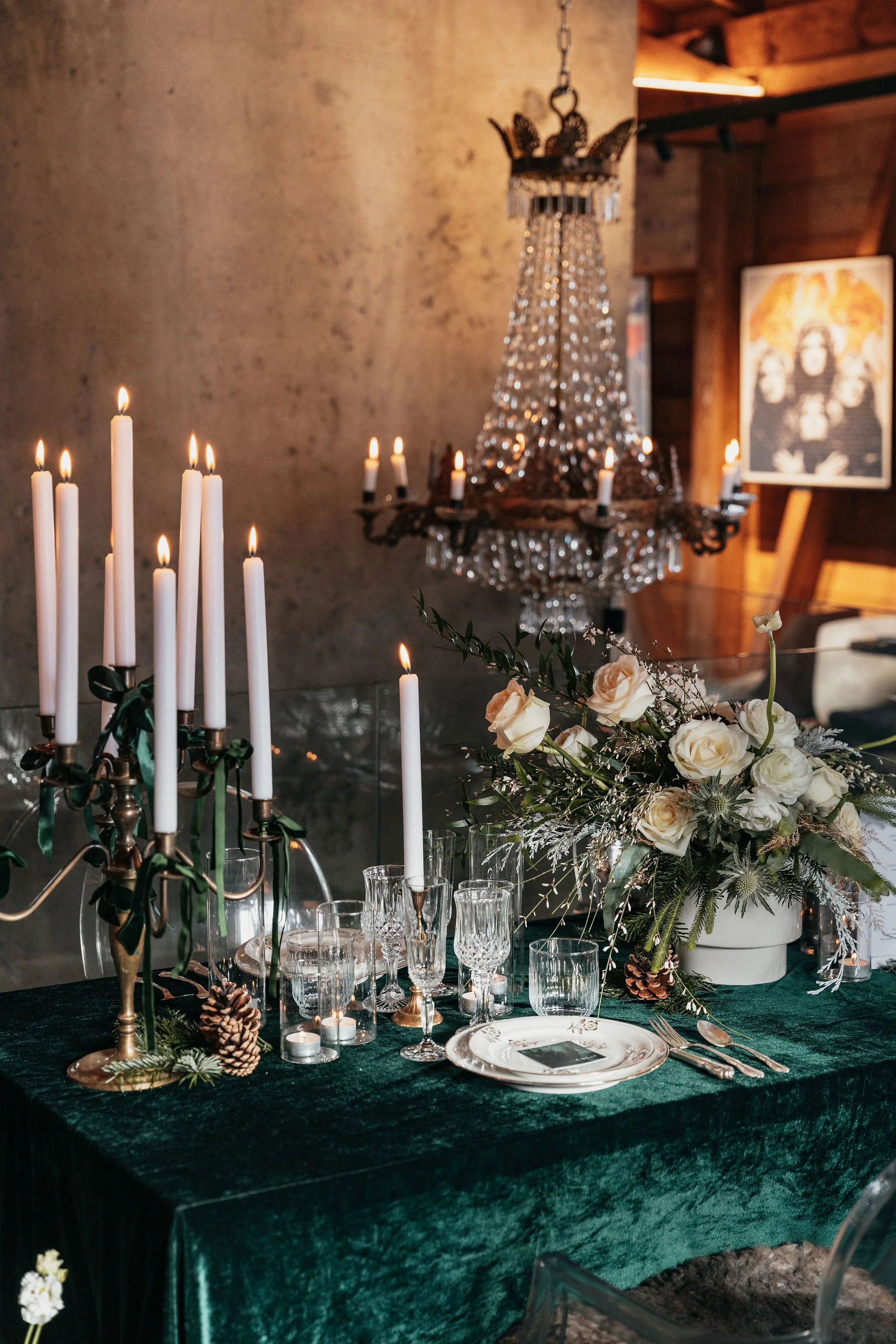 Table élégante décorée avec des bougies allumées, un arrangement floral blanc, des verres à vin, et une nappe en velours vert, dans une pièce chaleureuse avec un chandelier en cristal et une œuvre d'art en arrière-plan.