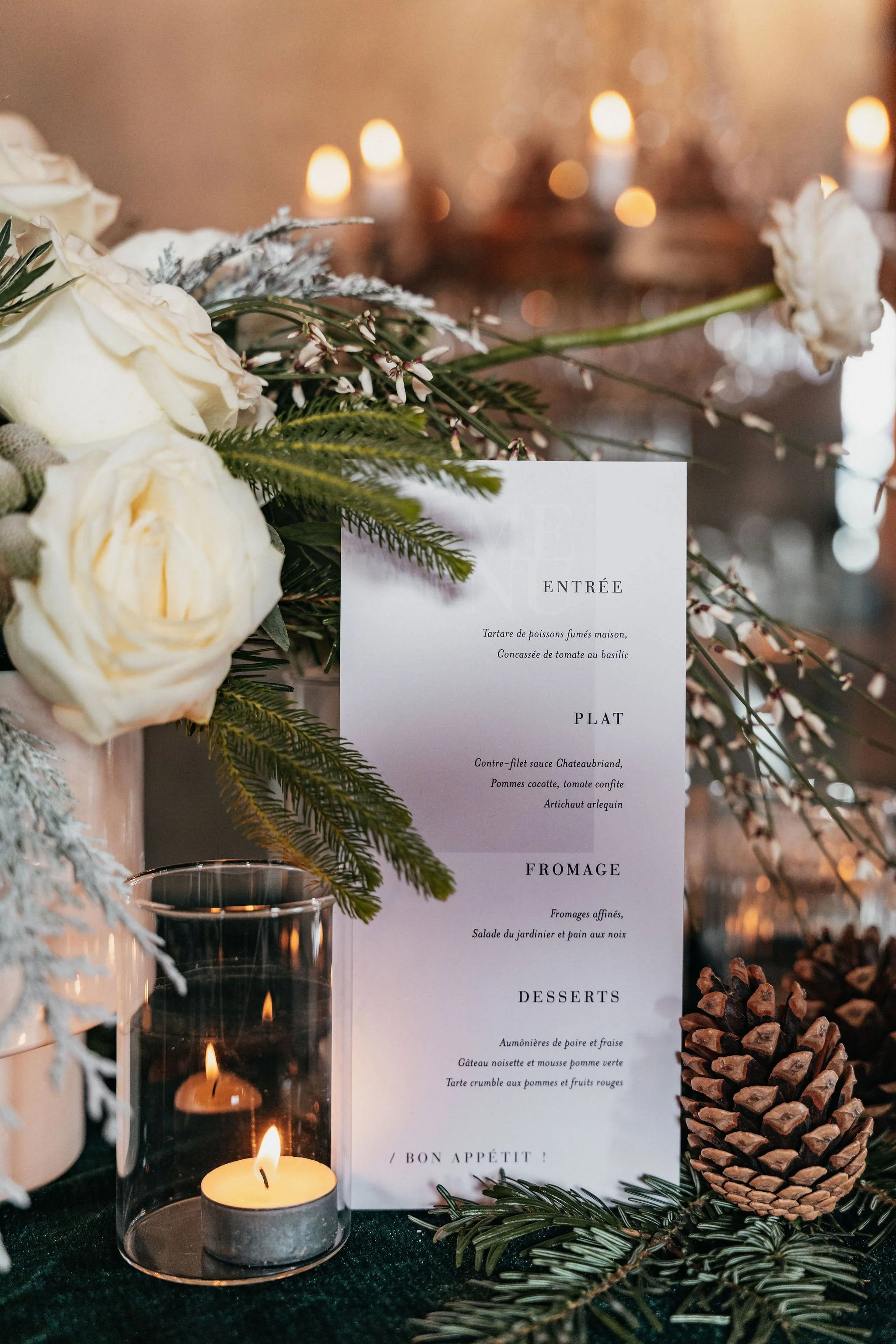 Un menu de restaurant placé à côté de bouquets de fleurs blanches, de branches de sapin et de pommes de pin, avec des bougies allumées en arrière-plan.