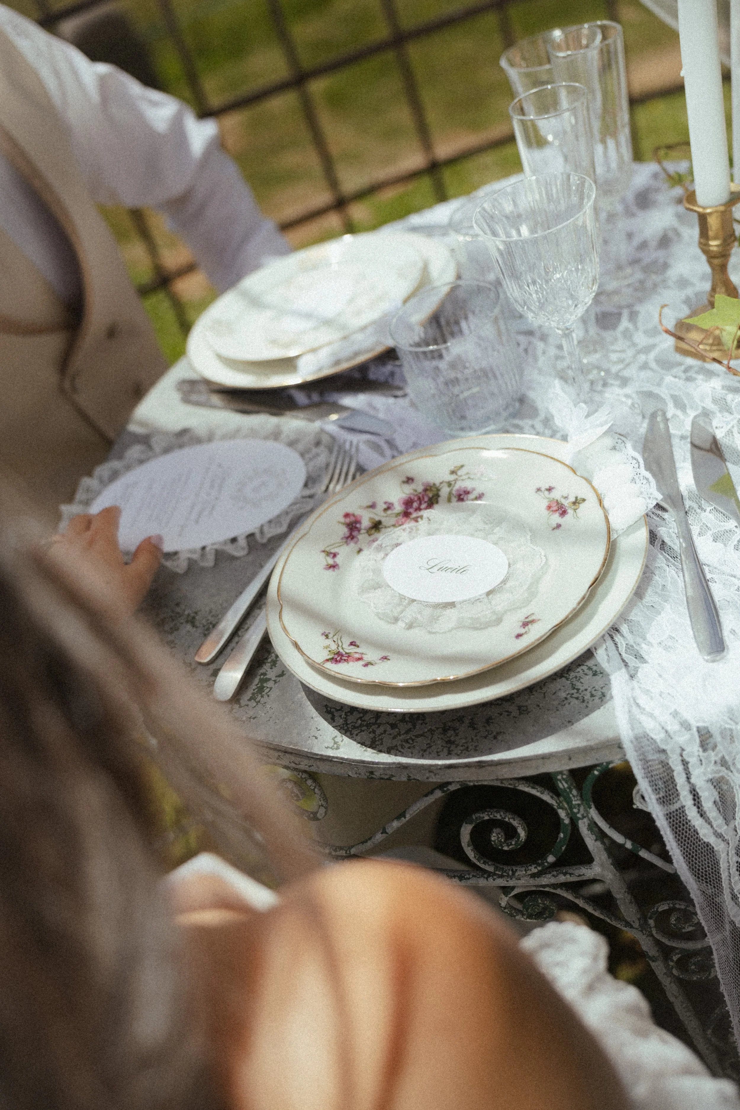 Table avec vaisselle en porcelaine à fleurs, quatre verres en verre, une bougie, et une nappe en dentelle blanche, à l'extérieur avec un décor de jardin.