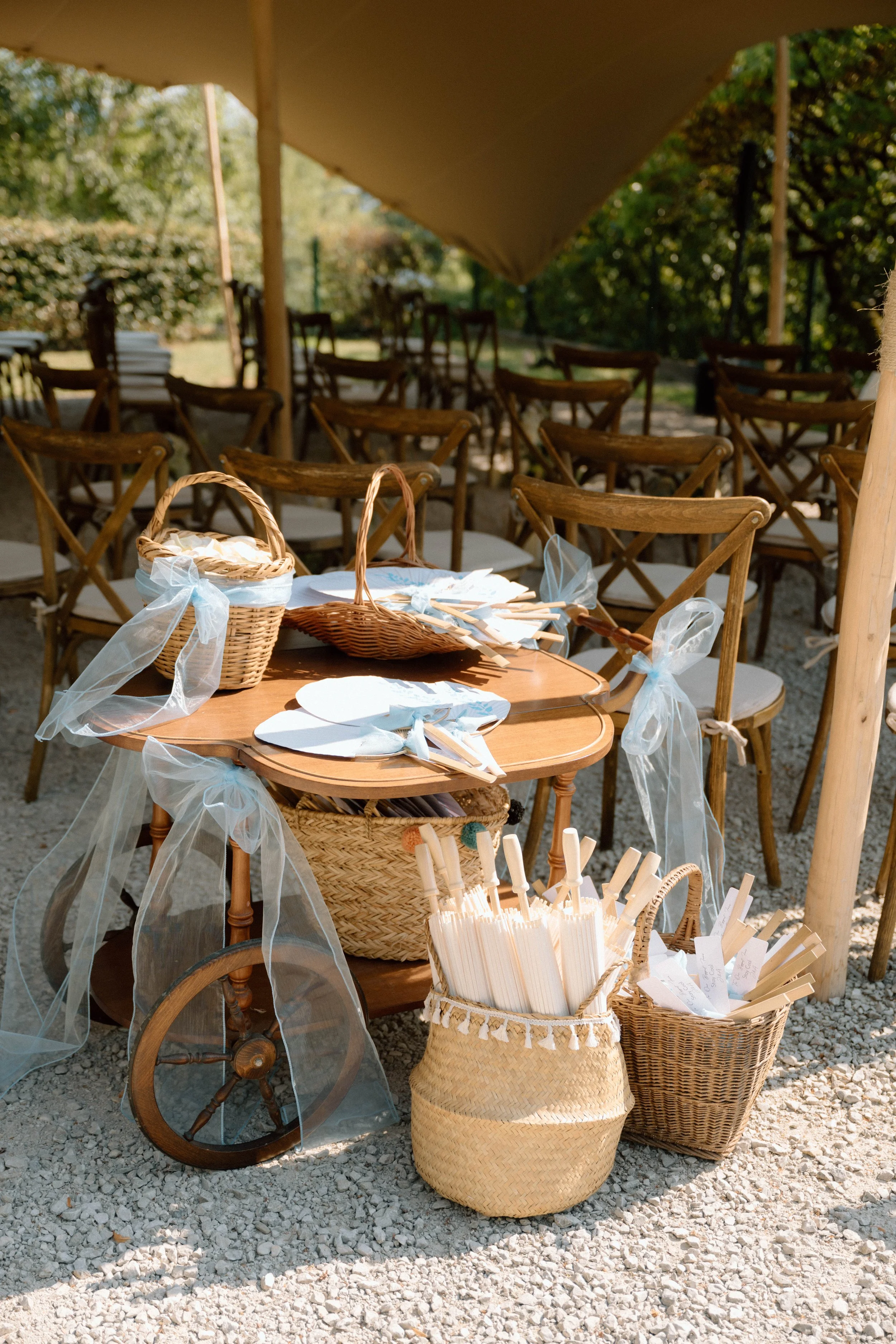 Table décorée avec des paniers contenant des serviettes en papier, des couverts en bois et des rubans en tissu blanc, entourée de chaises en bois décorées de rubans. En arrière-plan, un espace extérieur avec des chaises en bois et un parasol beige, s