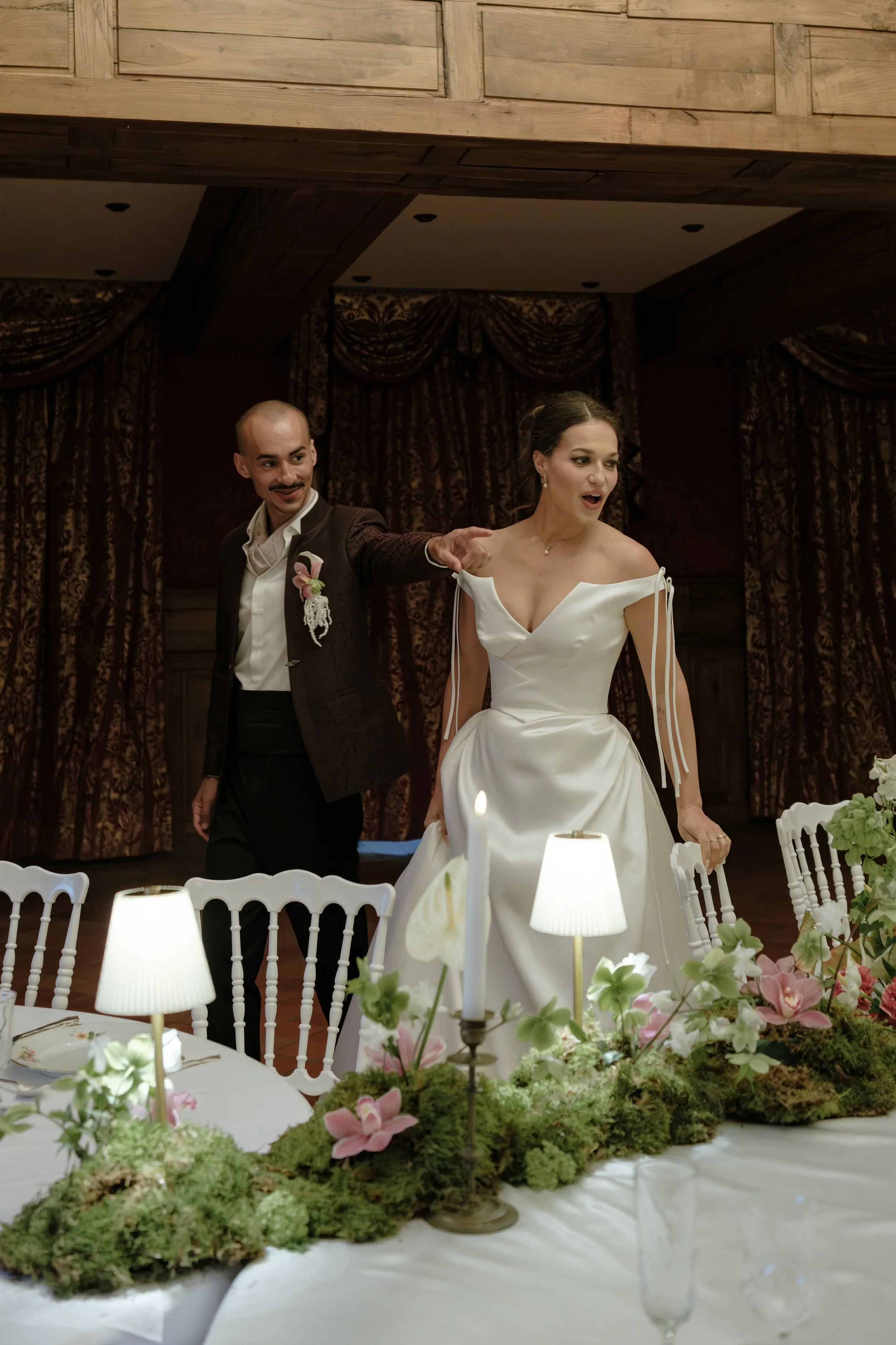 Un couple de mariage dans une salle décorée, avec un arrangement floral et des lampes sur une table. La femme porte une robe blanche et l'homme un costume sombre.