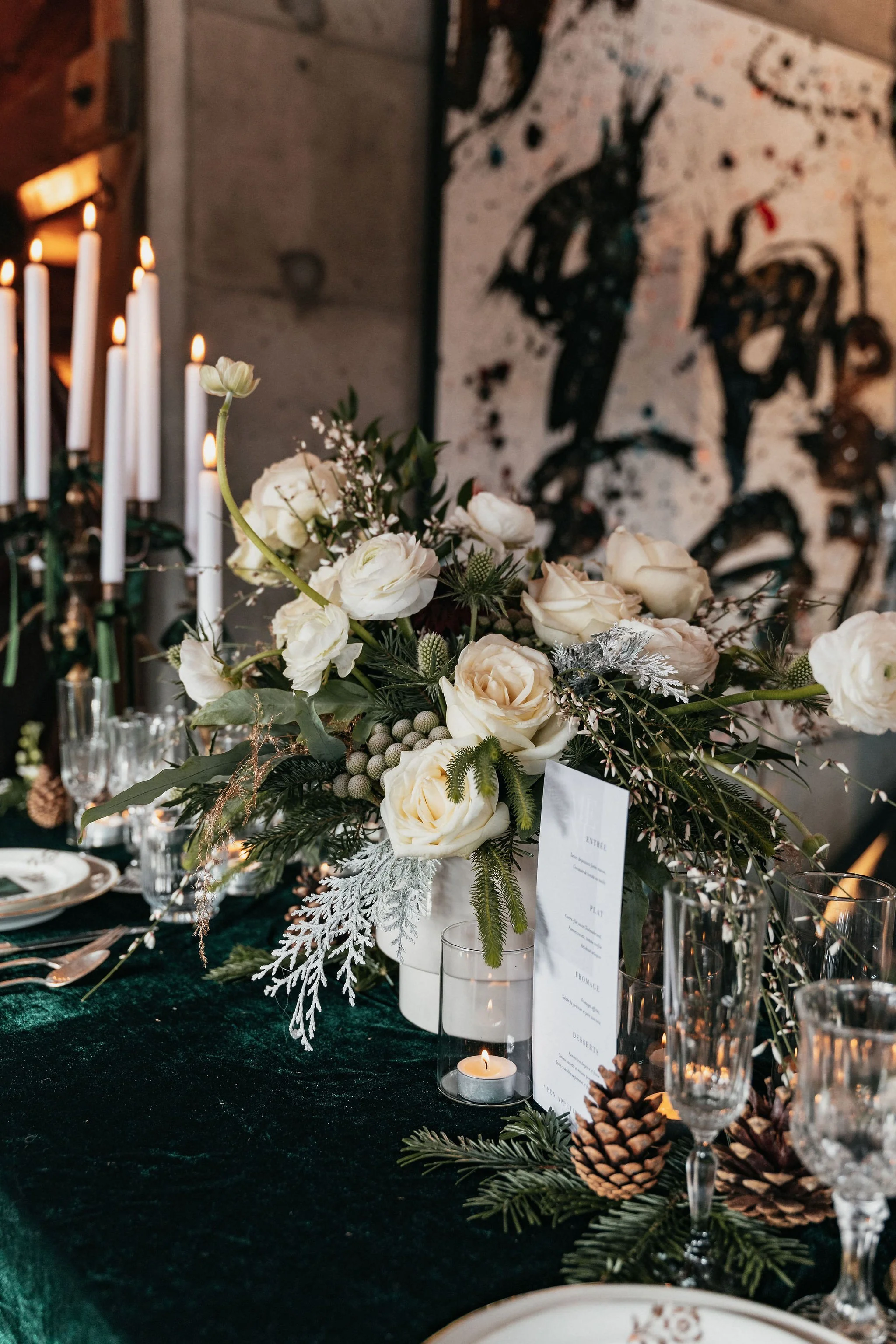 Une table de fête décorée avec un bouquet de fleurs blanches, des bougies et des éléments de décoration naturels comme des pommes de pin, dans une ambiance chaleureuse et élégante.