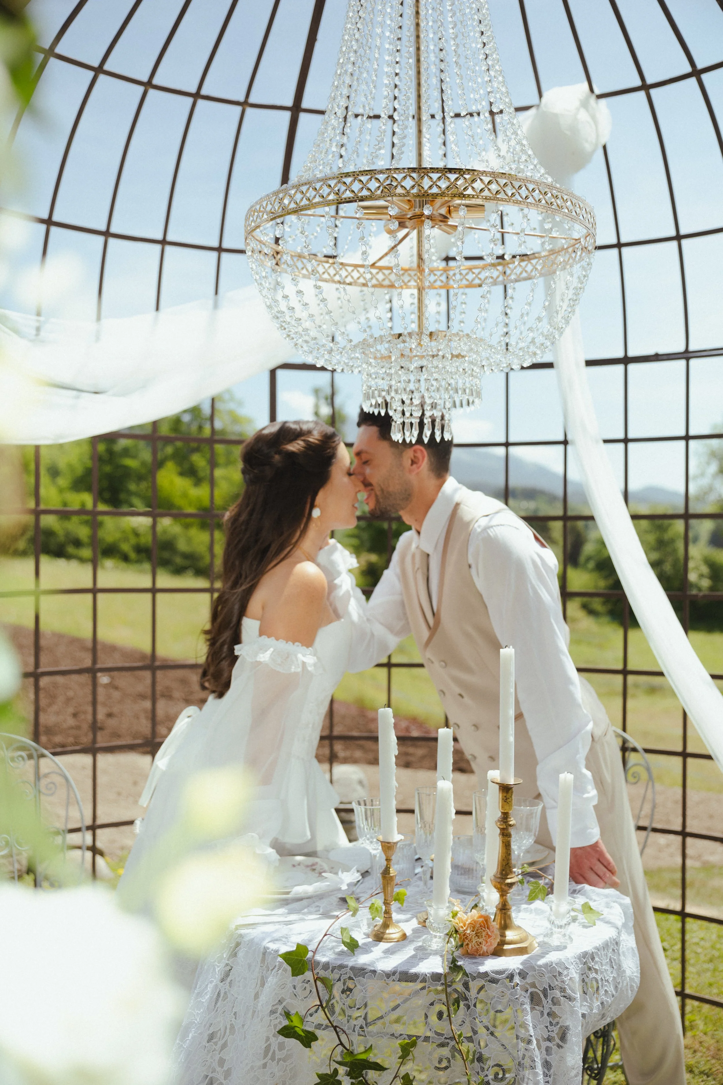 Un couple marié s'embrasse lors d'une cérémonie extérieure, sous une grande chandelier en cristal suspendu, avec une table ornée de bougies blanches et de décorations florales, dans un cadre en plein air avec un ciel bleu et une structure en treillis
